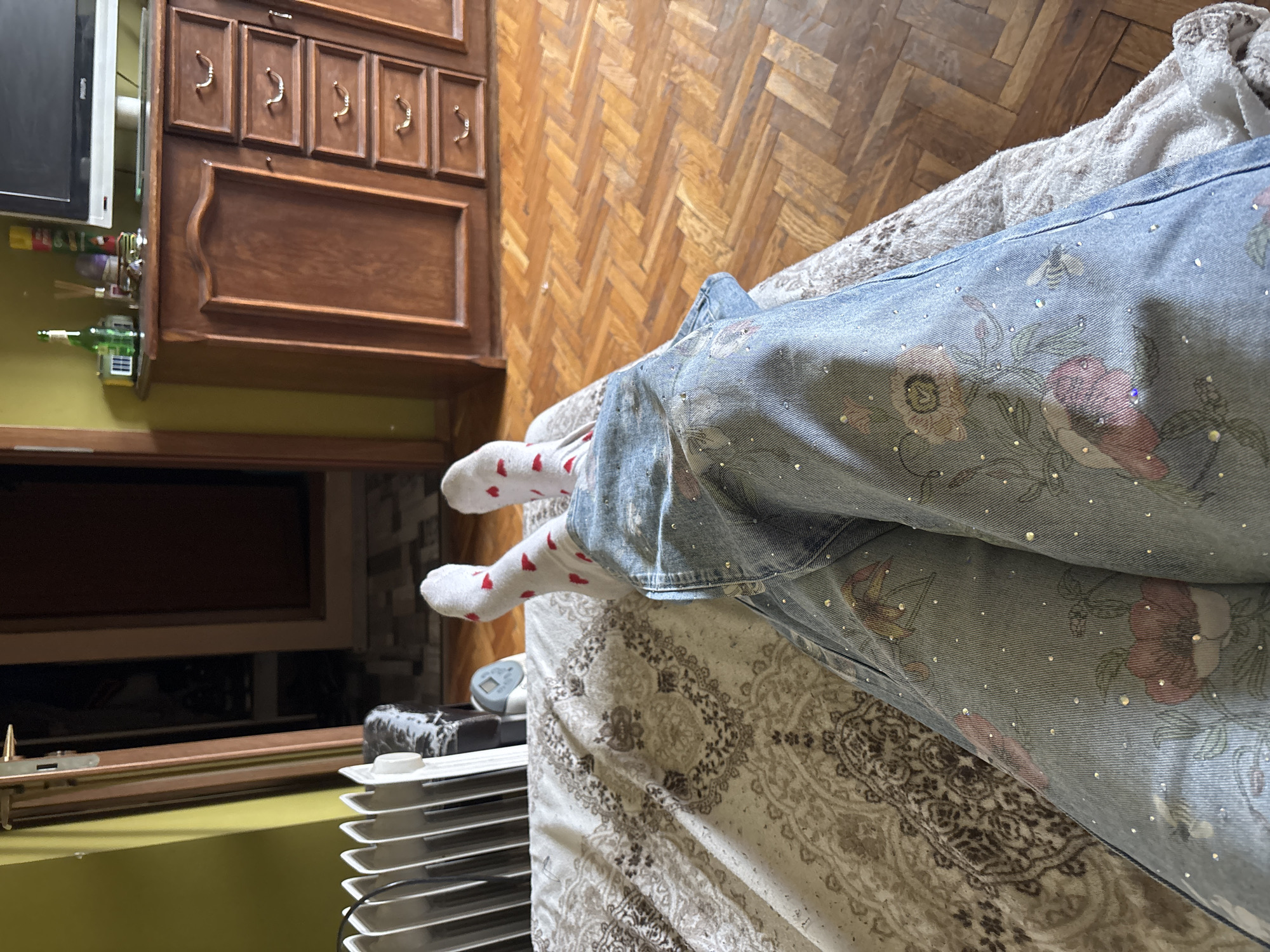 The image shows a pair of legs clad in light blue jeans with floral and sequin decorations, and white socks with red hearts. The person is lying on a bed with a patterned bedspread. The floor is parquet. In the background, there is a dark wooden cabinet and a radiator. The location appears to be indoors. It is not possible to confirm that the location is in Sofia, Bulgaria, based on the visual information provided.