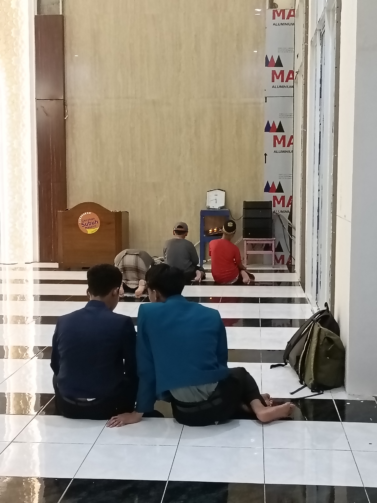 In Kediri City, Indonesia, an indoor scene captures four individuals engaging in prayer within a modern building. Two figures, seen from behind and dressed in dark blue and teal blazers respectively, are seated on the floor in the foreground. To the left, a wooden podium with an orange circular emblem and text reads "GERAKAN SUBH MANTRA" suggesting a spiritual or religious context. Further back, three men are in prayer postures on a tiled floor patterned with black and white squares. One man in a grey t-shirt and cap is kneeling, another in a checkered garment is prostrated with his head on the floor, and a third in a red t-shirt is seated. Various electronic devices, including speakers and a timer, are visible behind the praying individuals. The walls display vertical aluminum panels with repeating "MA" logos and abstract triangular designs. A backpack rests on the floor near the right edge of the frame. The lighting appears to be a mix of natural and artificial sources, illuminating the tiled floor and the figures.