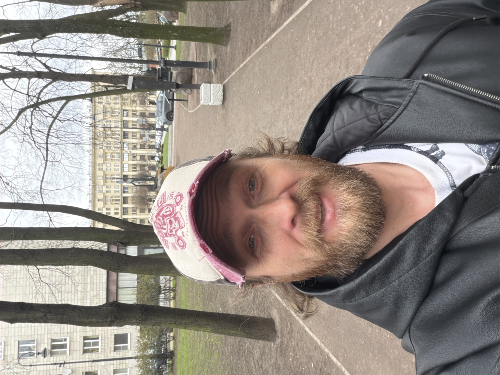 An outdoor, rotated selfie depicts a man with a reddish-blonde beard and long, light brown hair, smiling slightly at the camera. He has light green-blue eyes and is wearing a distressed white and pink baseball cap with a graphic design. The cap features the words "LIVE" and "CALIFORNIA," along with a stylized emblem resembling a skull or helmeted figure with goggles, and possibly the word "SINCE." He is dressed in a black leather jacket over a white long-sleeved t-shirt, which has a dark graphic print partially visible on the chest.
The background reveals an urban park or promenade, characterized by bare, deciduous trees suggesting late autumn, winter, or early spring. A paved path runs through the scene, flanked by grass or dirt areas. In the middle ground, tall, dark lamp posts are visible. Further back, there are multi-story buildings, including a prominent light-colored, ornate building with many windows, consistent with architecture found in Saint Petersburg, Russia. Other buildings and parked cars are also discernible. The sky is overcast, indicating a cloudy day. A small, white block-like object sits on the path in the distance. The man appears to be taking a casual photograph while enjoying a stroll.