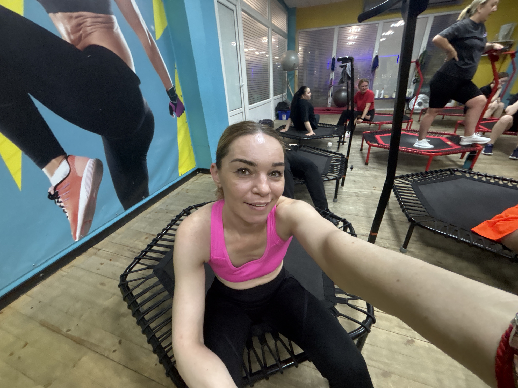 In a fitness studio, possibly located in Saint Petersburg, Russia, several individuals are engaged in a trampoline fitness class. The foreground features a woman with blonde hair, wearing a pink sports bra and black leggings, sitting on a black trampoline. She is smiling and holding the camera out to take a selfie. Behind her, other participants can be seen on their respective trampolines. To the left, a large mural depicts a woman in black athletic wear. In the background, there are exercise balls, gym equipment, and large windows. The floor is made of light-colored wood. The lighting suggests an indoor environment, likely a commercial gym.