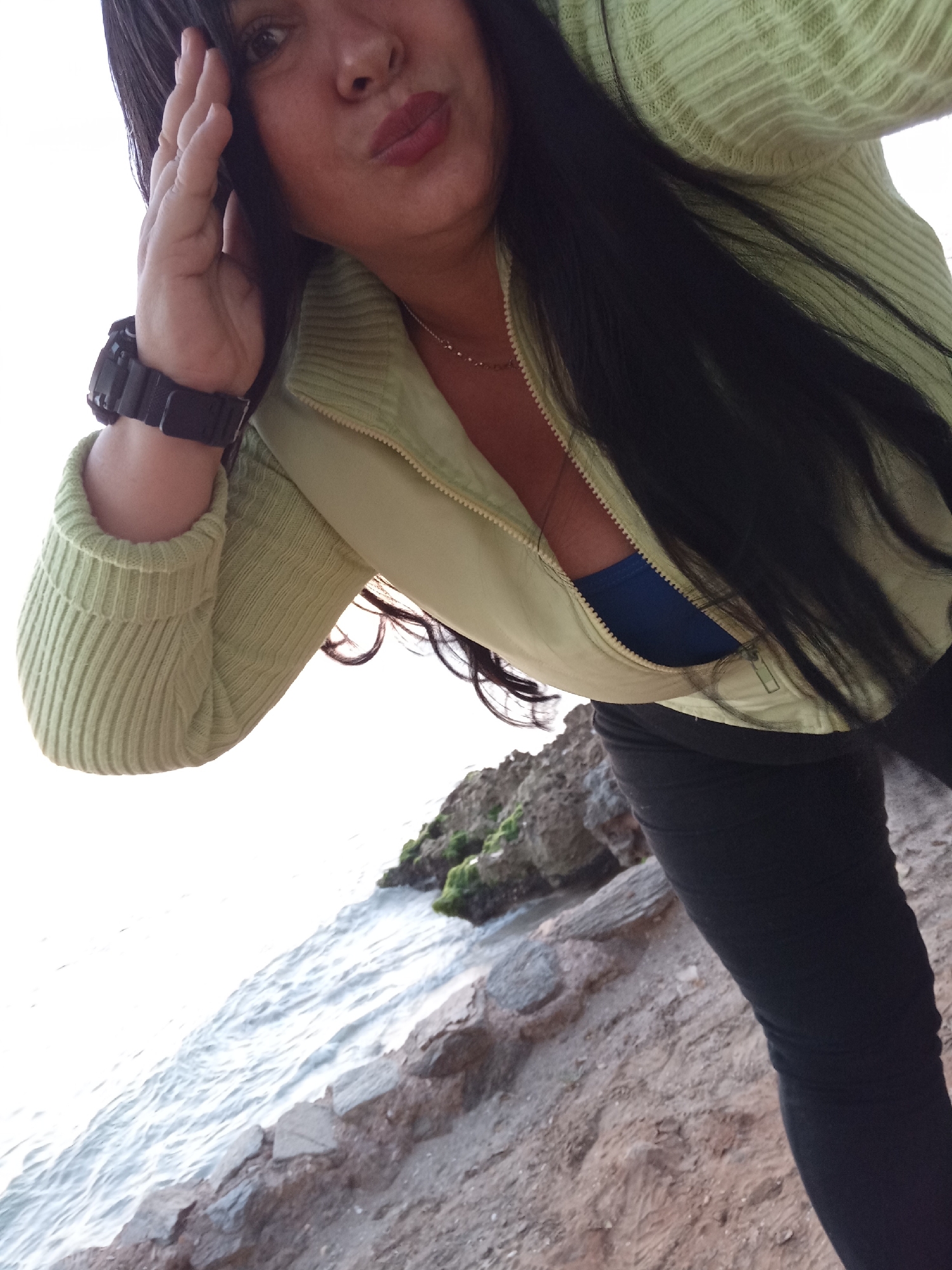 A medium-shot, selfie-style photograph taken outdoors shows a woman with long, dark hair and fair skin standing on a rocky beach, likely near the ocean.
The woman is wearing a light green zip-up sweater over a blue top and black leggings. She has a dark-colored wristwatch on her left wrist and a necklace. Her left hand is raised near her face. Her lips are pursed.
The foreground shows the beach with small rocks and what appears to be sand. In the background, to the left, is a body of water, and to the right, some larger rocks with some green vegetation growing on them.
Based on the prompt indicating that this photo was taken in the city of Juan Griego, Venezuela, it can be inferred that this is likely a coastal area in that region.