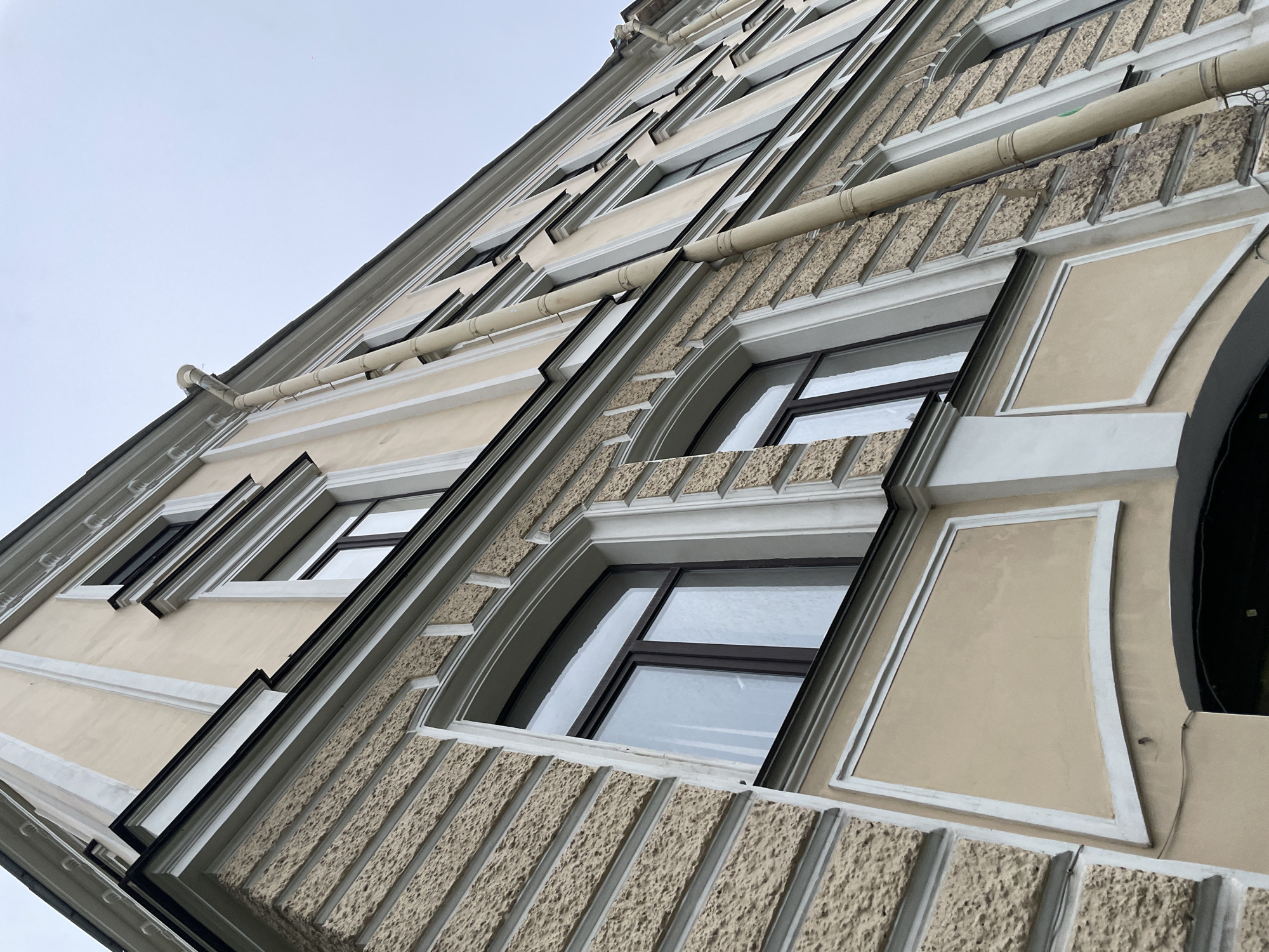 The image depicts the exterior of a building in Saint Petersburg, Russia, viewed from a low angle. The facade features a combination of beige and textured stone-like surfaces, with windows set within arched and rectangular frames. A light-colored pipe runs horizontally along the building's facade. The sky is overcast. Architectural details such as decorative moldings and window styles suggest a historical or classical design.