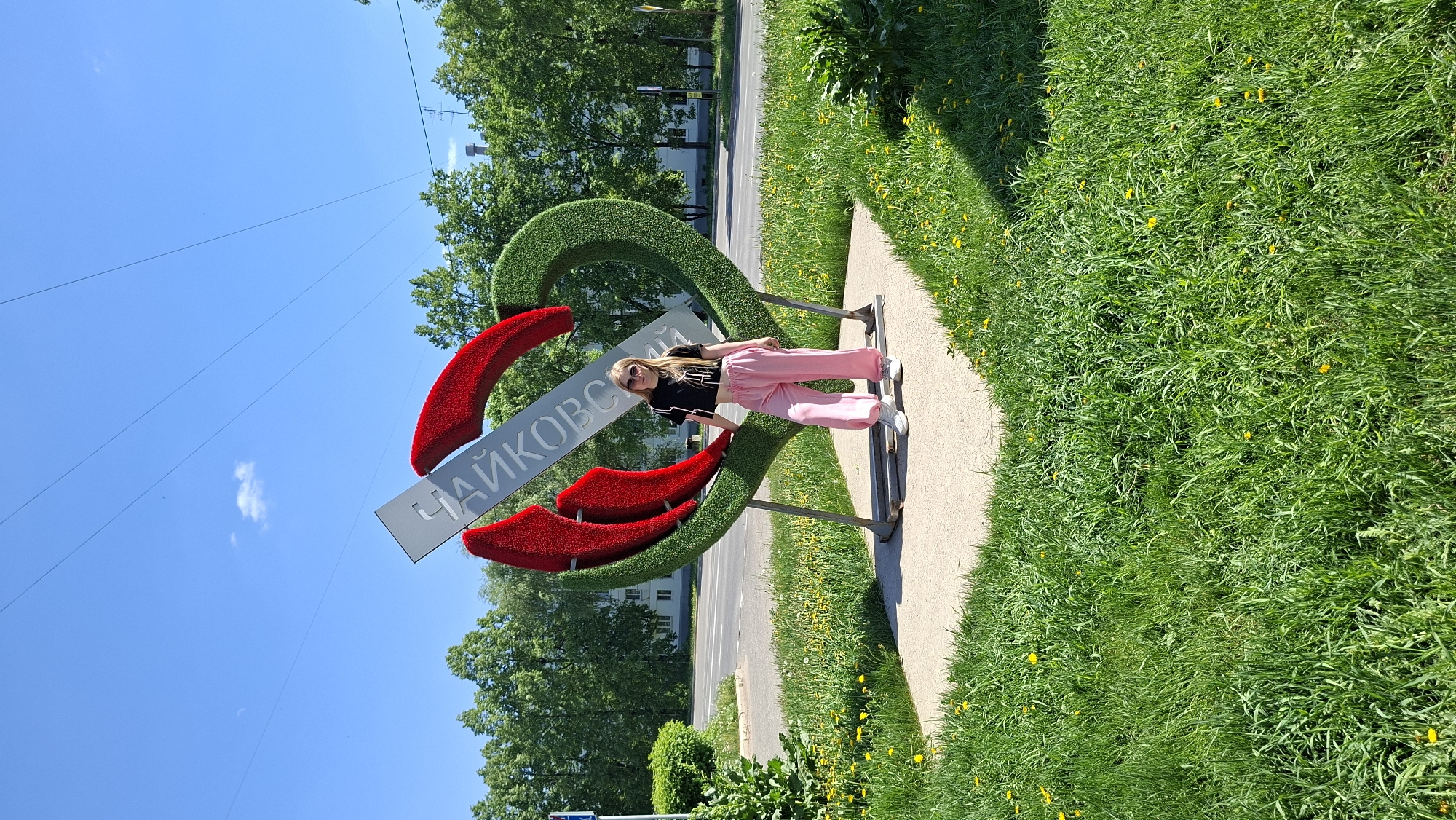 A fair-skinned young woman with long blonde hair is standing on a concrete walkway next to a large, three-dimensional, artistic sign. The sign is shaped like an abstract flower or emblem, with a green outer outline and red accents. A horizontal silver plaque displays the name "Чайковский" (Chaikovsky) in Cyrillic script. The woman is wearing a black top and pink pants, with white sneakers. The scene takes place on a sunny day, with a blue sky visible in the upper left corner. Green grass and trees surround the sign, indicating it is in a park or outdoor area. A road is visible in the background. The location is likely the city of Chaikovsky, Russia, based on the Cyrillic inscription on the sign.