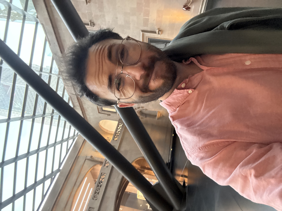 A male subject, approximately 30-40 years old, with dark, disheveled hair, a full beard, and gold-rimmed aviator-style glasses, poses for a selfie. He wears a light pink collared button-down shirt with the top buttons undone and has a dark green or grey jacket draped over his left shoulder. His expression is a slight, amused smirk, with his left eye appearing more squinted than his right.
The scene is set indoors, within the grand, multi-story interior of a historical building in the Historic Center of Mexico City, Mexico, specifically characteristic of the **Palacio de Cultura Citibanamex - Palacio de Iturbide**. The background features light-colored stone walls exhibiting colonial or neoclassical architecture, including multiple arched openings on an upper level and ornate, decorative balustrades. The space is covered by a modern, gridded glass roof, through which blurred reflections or distant views of contemporary high-rise buildings are visible. Large, dark, diagonal tubular structures, possibly architectural supports or an art installation, traverse the upper left portion of the frame. The floor in the midground appears dark and reflective. The lighting is bright, predominantly natural light entering through the glass roof.
