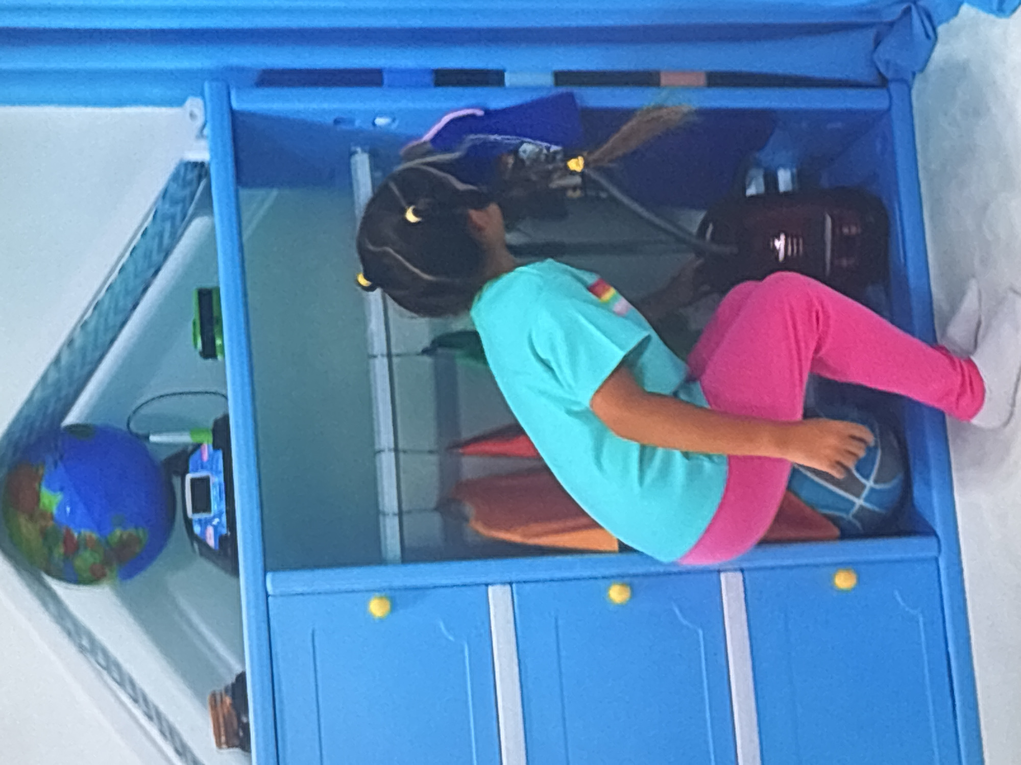 The image shows a young girl sitting inside a blue plastic cabinet or toy storage unit. She is wearing a teal t-shirt and pink leggings. Her hair is dark and styled with yellow ties. In the cabinet, there are various objects including a blue and green globe, what appears to be a tablet or small screen device, and a dark brown object that could be a violin case. There are also some orange and red items that are difficult to identify clearly. The girl is holding a blue basketball in her lap. The cabinet is blue and has yellow knobs on its doors. The scene appears to be indoors, likely a child's room. There is no visible text in the image. The lighting suggests it is daytime. The context provided indicates the location is Dubai, United Arab Emirates.