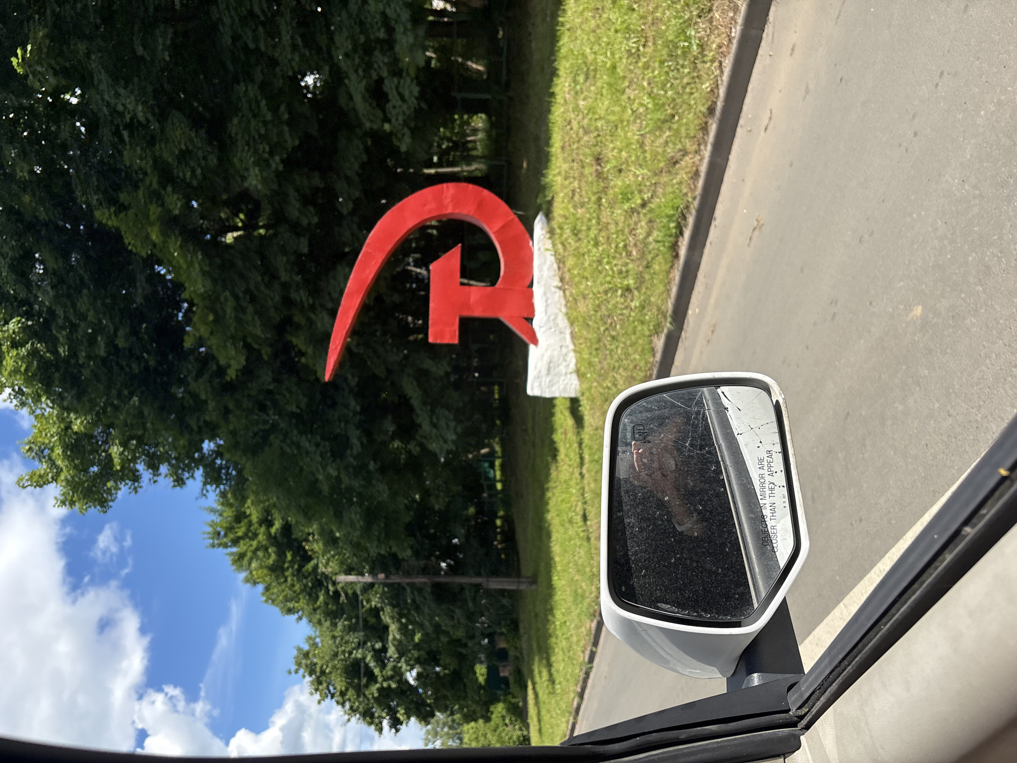 A red hammer and sickle sculpture is visible on a grassy mound next to a road. The sculpture, a symbol of communism, is large and metallic, with the hammer and sickle intertwined. The base of the sculpture is white and appears to be concrete. The road is asphalt and runs alongside the grassy area. In the foreground, a car's side mirror is visible, reflecting a person's hand holding a phone. The mirror has text that reads "OBJECTS IN MIRROR ARE CLOSER THAN THEY APPEAR". The background consists of lush green trees and a blue sky with white clouds. The scene appears to be in Pereslavl-Zalessky, Russia, a city known for its historical significance and Soviet-era monuments.
