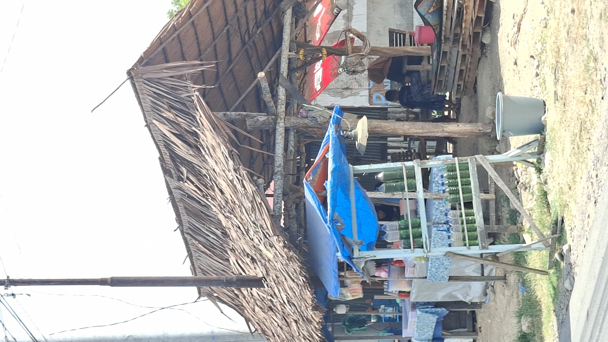 The image depicts a roadside stall in Lhokseumawe, Indonesia, under a large, weathered thatched roof. The stall is selling bottled green liquid, possibly fuel, displayed on a wooden rack in front. Various other items are visible on tables covered with blue and white patterned cloths. Behind the stall, a few people are present, including someone sitting in the shade and a child or young person facing away from the camera. The structure appears rustic, with exposed wooden beams and a corrugated metal roof underneath the thatch. The ground is dirt and gravel, with some sparse vegetation. The overall impression is of a simple, functional roadside business operating in a warm, sunny climate.