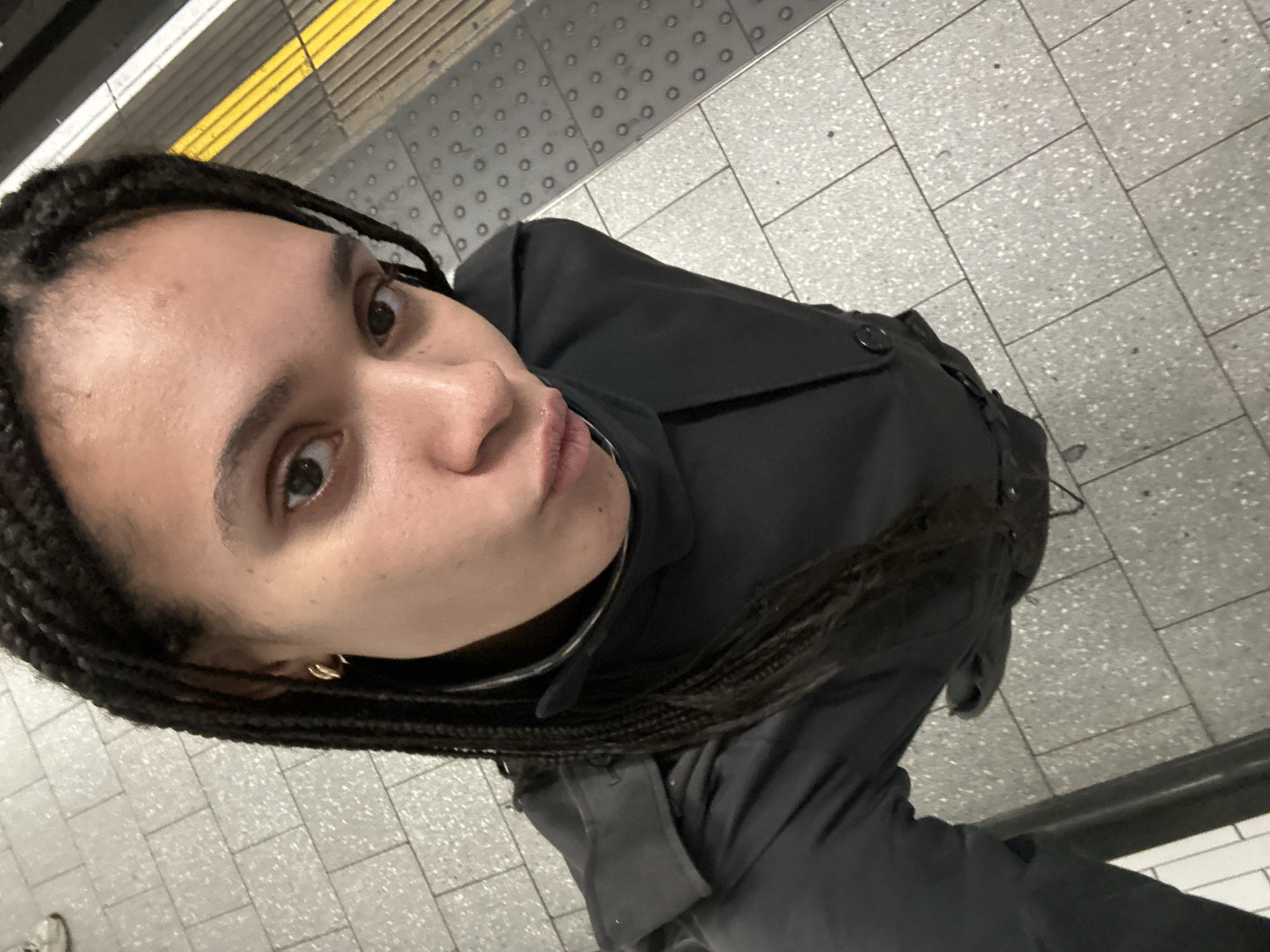A woman with braided hair takes a selfie in a London Underground station. She is wearing a dark coat. The platform floor is tiled in grey and white. A yellow safety line runs along the platform edge. A metal handrail is visible in the lower right corner.