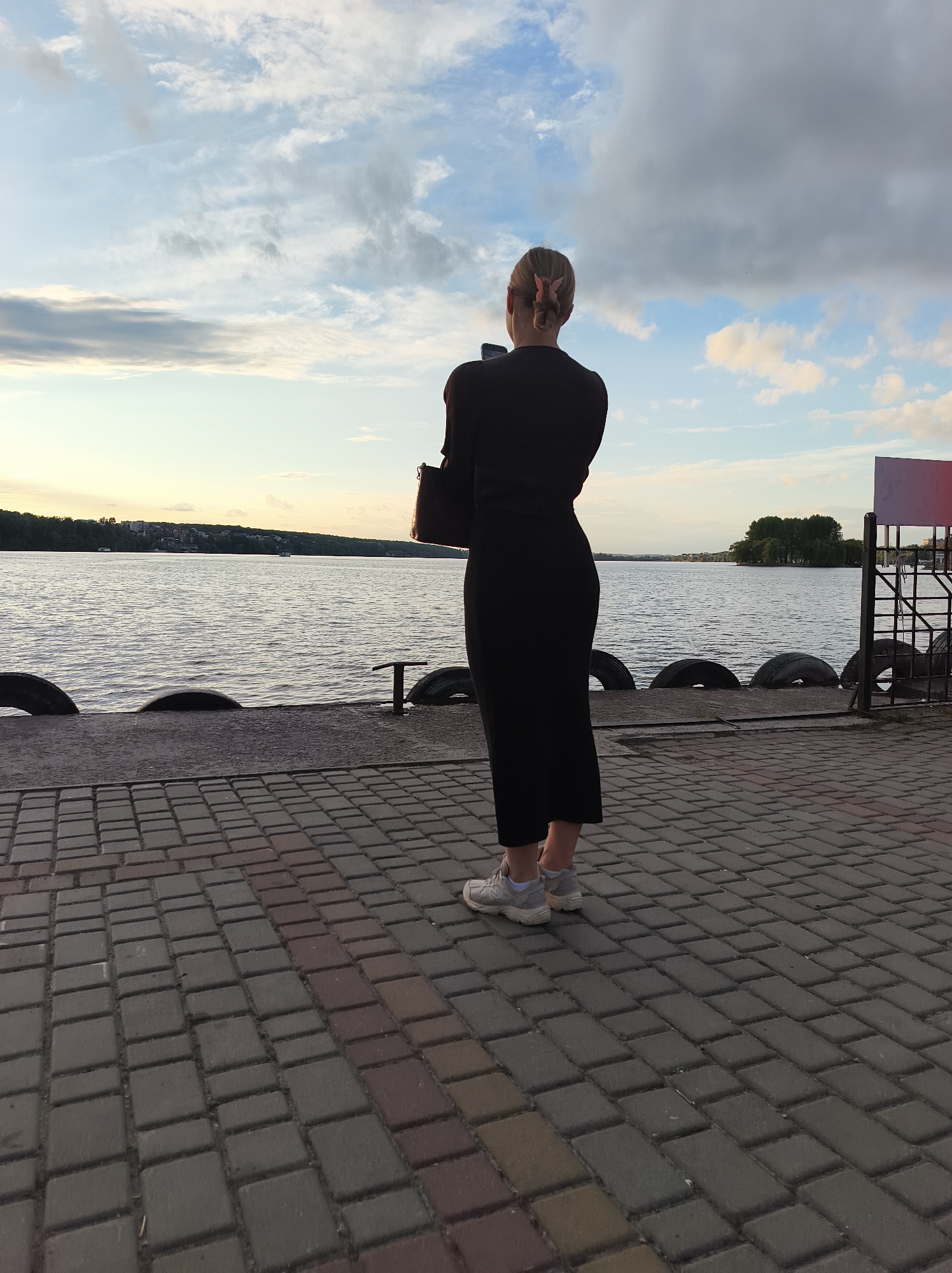 A woman stands on a paved pier overlooking a body of water. She is facing away from the camera, holding a smartphone in her right hand. She is wearing a black, long-sleeved dress and white sneakers. Her hair is pulled back in a bun and adorned with a pink clip. The pier is made of interlocking pavers. Along the edge of the pier are large, black tires used as fenders. In the background, the water is calm, and a tree-covered island is visible in the distance. The sky is partly cloudy with a soft, warm light, suggesting either sunrise or sunset. The scene appears to be the Ternopil lake, a large man-made reservoir within the city of Ternopil, Ukraine.