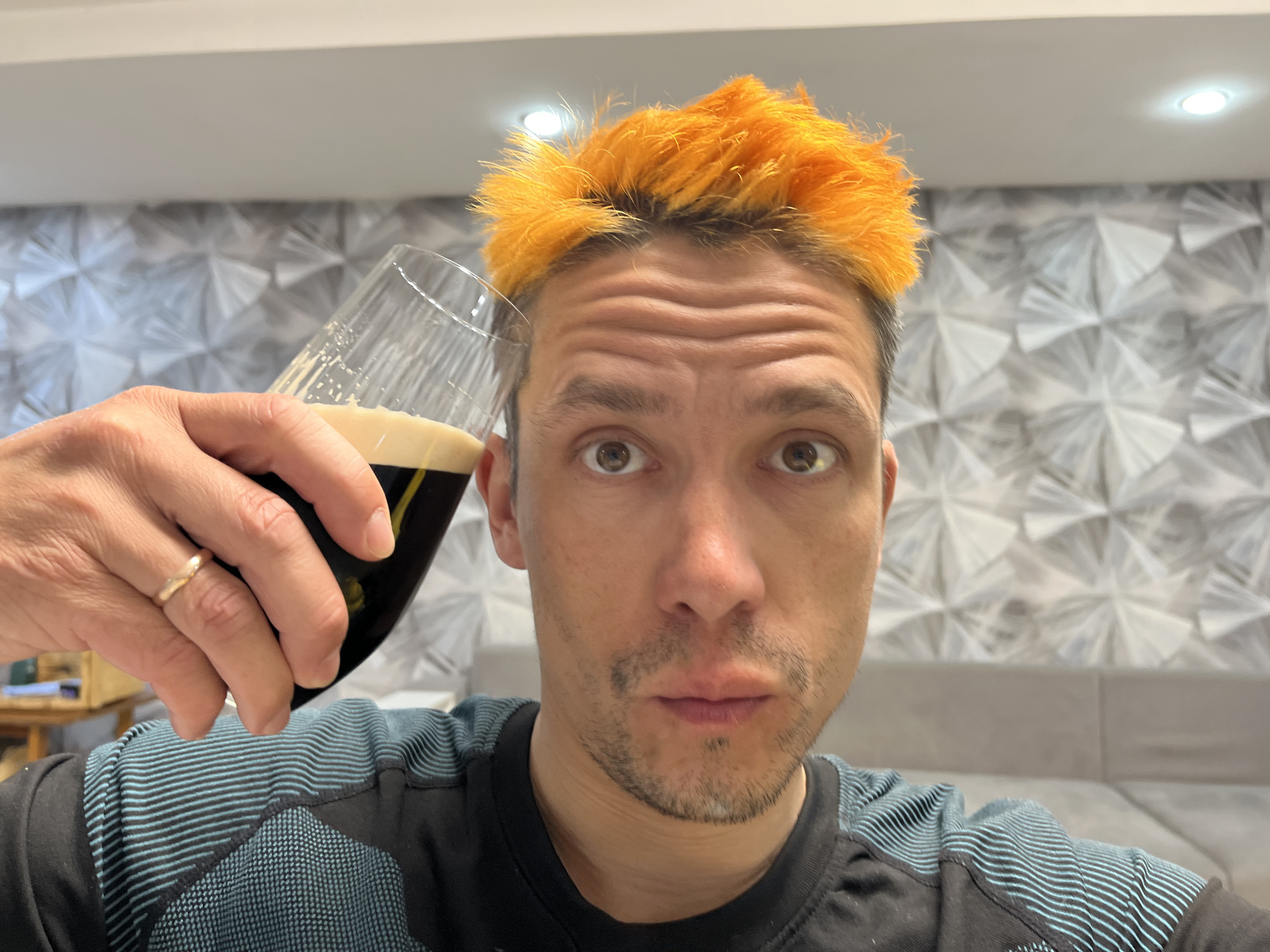A man with short, bright orange, spiky hair and light stubble stares wide-eyed at the camera in a close-up, indoor selfie. His eyebrows are raised, creating horizontal wrinkles on his forehead, and his lips are slightly parted, giving him a surprised or inquisitive expression. He wears a black long-sleeved shirt with light blue horizontal stripes across the shoulders and upper arms.
In his left hand, on which a gold-colored ring is visible on his ring finger, he holds a stemmed glass filled with a dark liquid, likely a stout or dark beer, topped with a creamy, light-colored foam head. The glass is tilted, indicating it might have just been lifted or is about to be consumed.
The background shows a modern interior, consistent with a residential setting in Chaikovsky, Russia. Behind the man, a textured wall features a striking geometric pattern of light grey and white facets. Above this patterned wall, a smooth, light-colored ceiling is visible, featuring a single recessed light fixture providing illumination. A portion of a light grey upholstered couch or sofa can be seen in the lower right background. The artificial lighting suggests the photo was taken indoors, possibly in the evening. There is no visible text in the image.