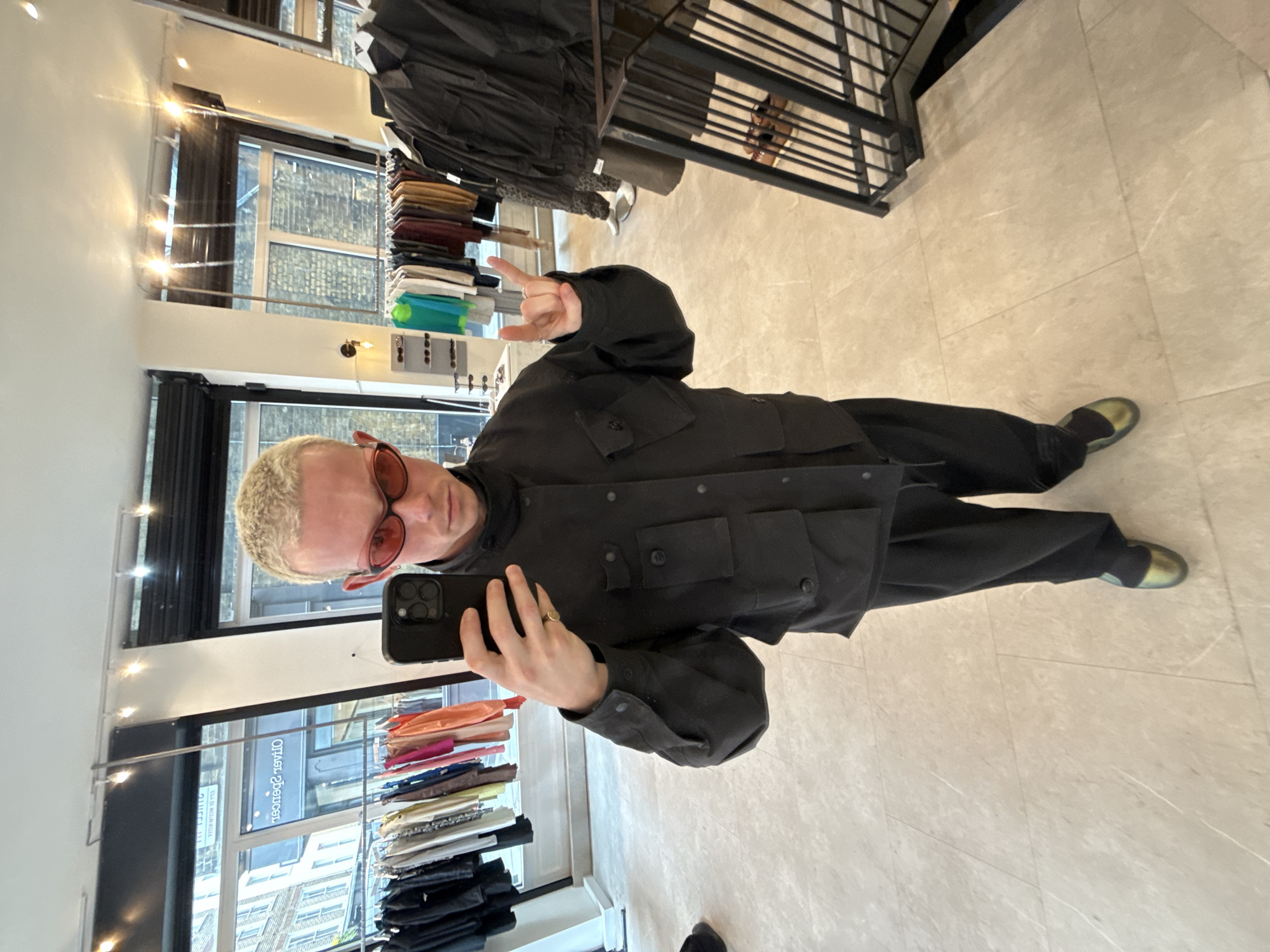 The image depicts a person taking a selfie in what appears to be a clothing store in London, United Kingdom. The person has short, bleached blonde hair and is wearing red sunglasses, a black jacket with pockets, and black trousers. They are holding a black smartphone in their left hand, presumably to take the picture, and making a hand gesture with their right hand. The store has clothing racks with various items, including garments in shades of brown, blue, and orange, displayed near windows. The background indicates a brick building visible through the window. The floor is tiled, and there are lighting fixtures visible, suggesting an interior setting.