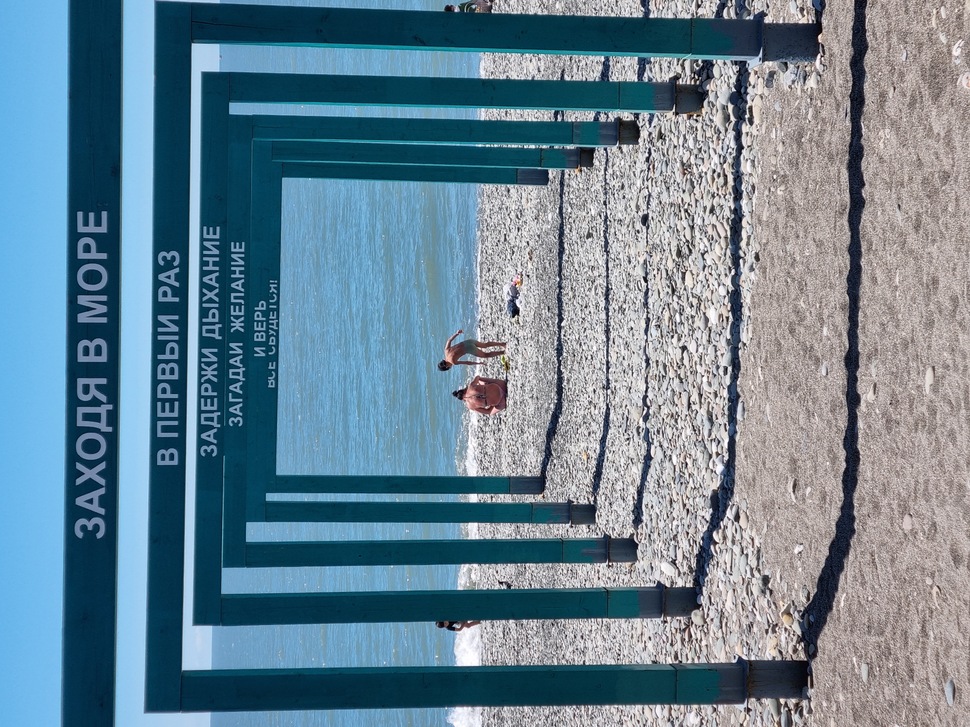 The image shows a view of a beach in Sochi, Russia, framed by a series of teal-colored wooden structures with text in Russian. The text, which appears on the left side of the frames, reads "Заходя в море", "В первый раз", "Задержи дыхание", "Загадай желание", "И верь", "Все сбудется!". The beach is composed of small pebbles and sand. In the center of the frame, people are visible on the beach and in the water. A child stands near the water, and an adult is sitting on the beach. The sea is calm with small waves. The scene is bathed in bright sunlight, casting shadows of the wooden structures across the beach.