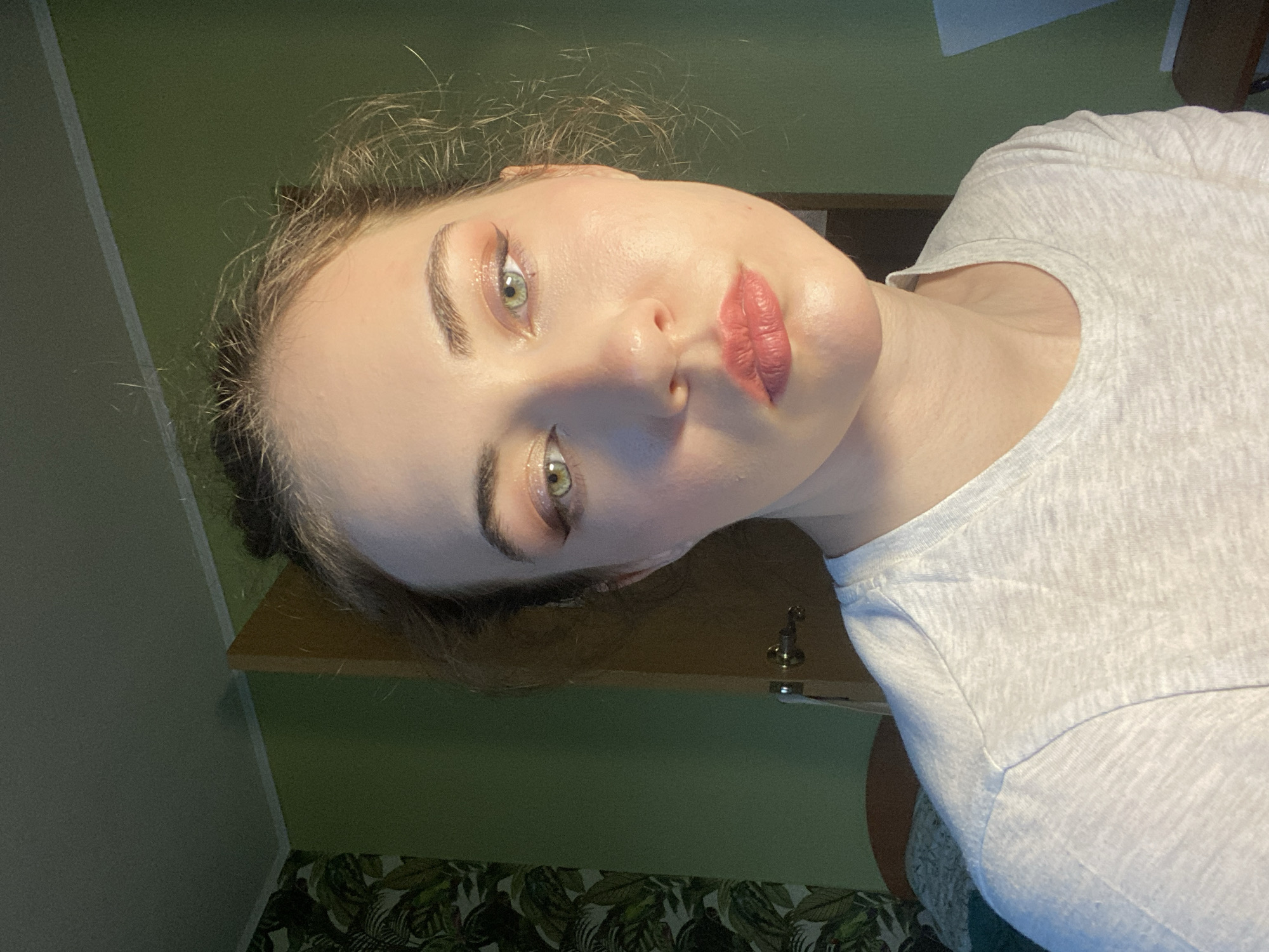 This is a close-up, vertically oriented, eye-level shot of a young woman's face, tilted to the side. She is wearing a light gray t-shirt and has her hair pulled up loosely, with some strands framing her face. Her makeup includes eyeshadow and lipstick in muted pink and coral tones. She has striking green eyes, and her skin appears smooth and lightly made up. The background is a plain green wall, with a wooden shelf and a sliver of a wooden drawer visible. A dark, leafy patterned wallpaper can be seen at the bottom of the frame. The lighting suggests an indoor setting, possibly artificial. The overall mood appears relaxed and candid. No visible text is present in the image.