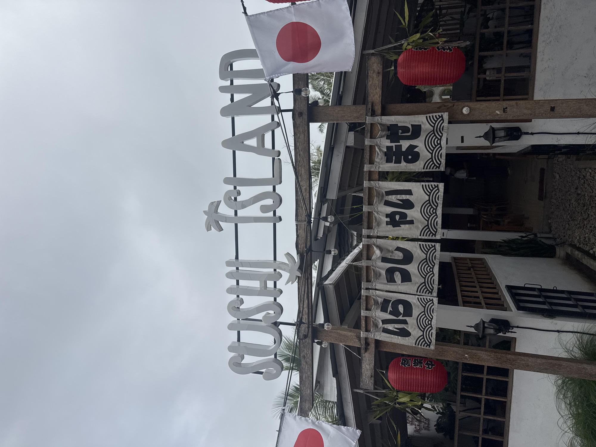 The image shows the exterior of a Japanese restaurant named "Sushi Island" located in Nusa Penida, Indonesia. The sign is made of large, white letters arranged vertically. Two Japanese flags and two red paper lanterns are attached to the wooden structure supporting the sign. Black and white fabric banners with Japanese script hang vertically from the same structure. The restaurant building is visible in the background, with white walls, windows, and decorative elements. The sky is overcast.