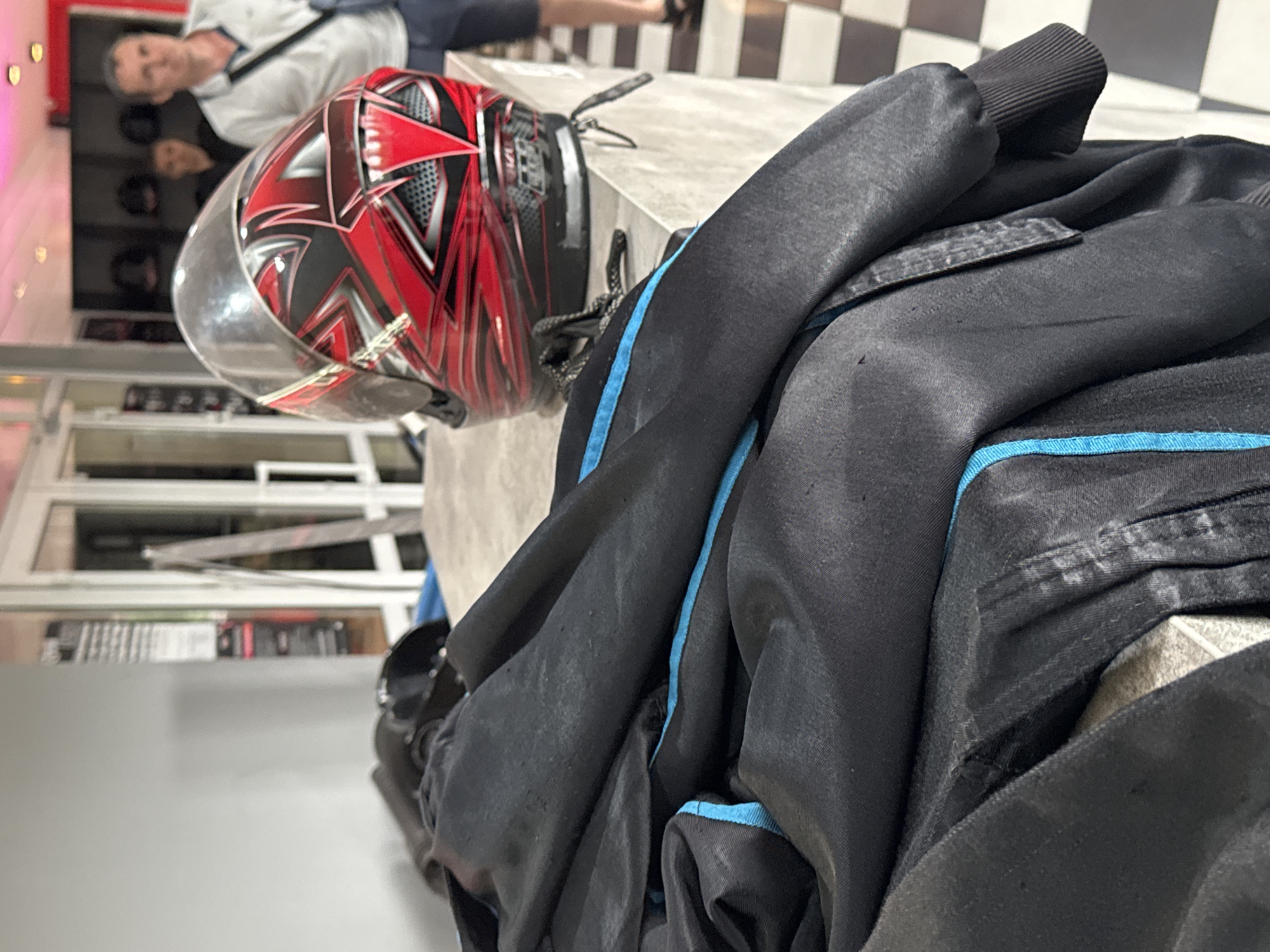 The image shows a red and black motorcycle helmet on a light-colored surface. A black jacket with blue trim is draped over the helmet. In the background, partially visible, are people and shop windows. The presence of people in the background suggests an indoor location in the city of Perm, Russia.