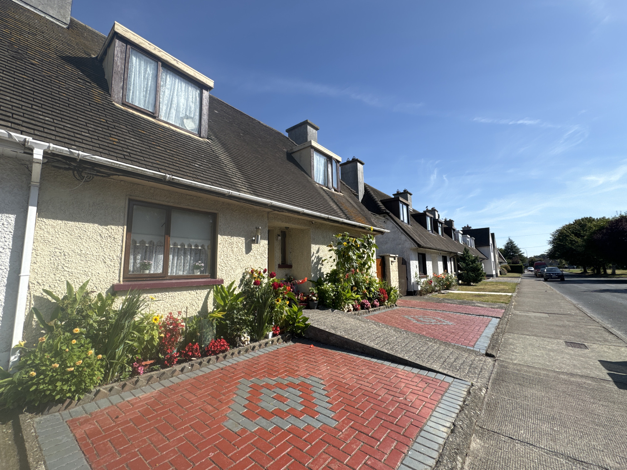 The image shows a row of terraced houses on a sunny day in Waterford, Ireland. The houses are light-colored with dark roofs and dormer windows. In front of the houses are gardens with colorful flowers and red brick driveways with gray patterns. A concrete sidewalk runs along the street, and cars are visible in the distance. The sky is a clear blue with some wispy clouds.