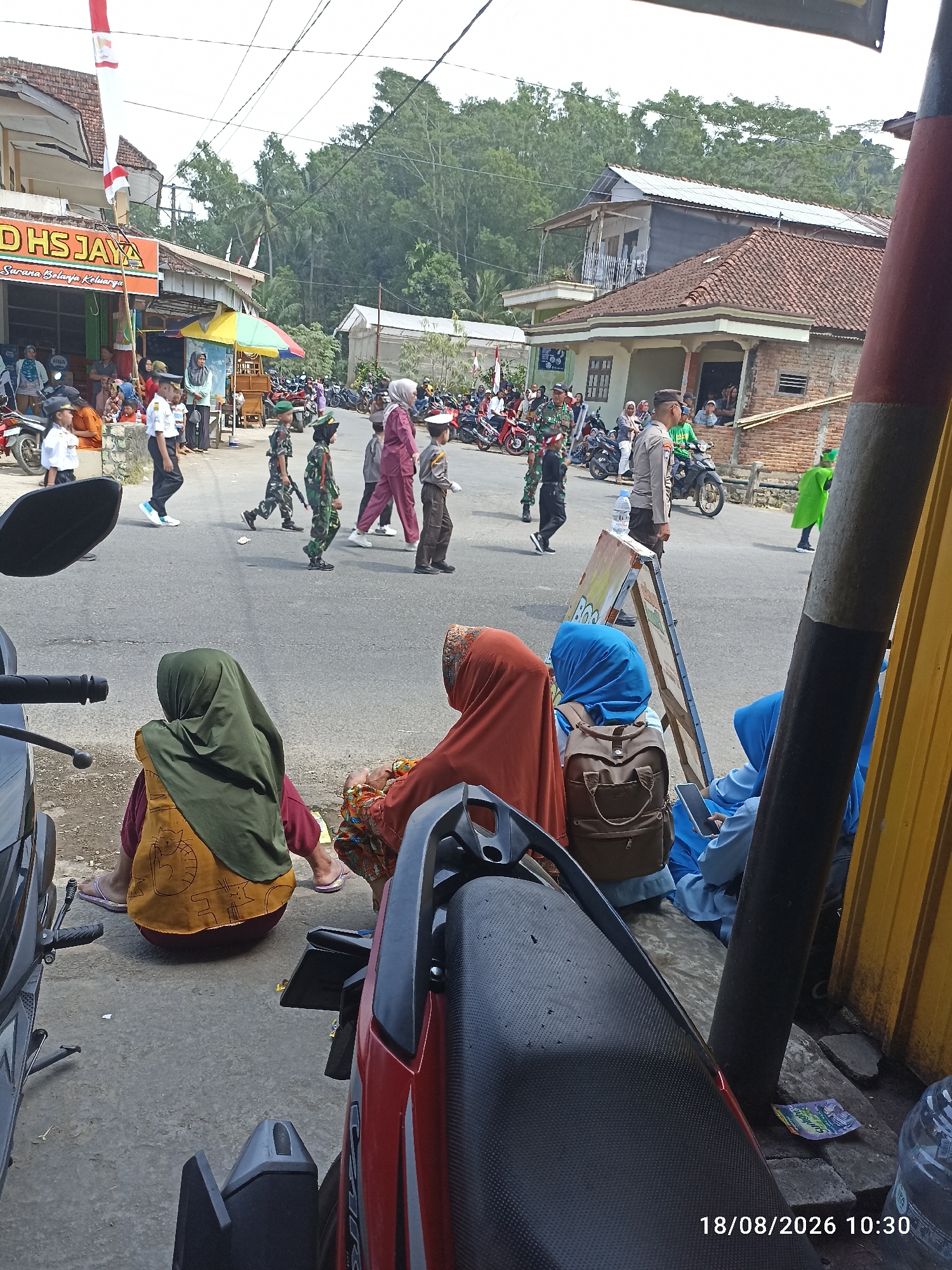 In a street scene in Unknown, Unknown, a parade appears to be in progress. Several children in uniform, some in military-style outfits and one in a pilot's uniform, walk in a procession on the asphalt road. Spectators are gathered on the side of the road, some seated on the ground. One person, possibly a police officer, stands on a motorcycle. The environment shows buildings lining the street, with trees and vegetation in the background. The day is bright, suggesting it is daytime. There are several motorcycles parked along the side of the road. In the foreground, parts of motorcycles are visible, and a yellow structure, possibly a fence or wall, is present on the right. A timestamp in the lower right corner indicates "18/08/2026 10:30".