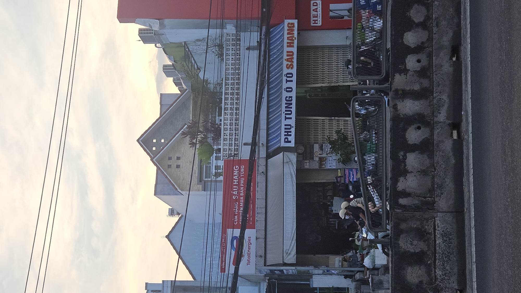 The image captures a street scene in Cam Đức, Vietnam, featuring storefronts along a roadway. The building on the left has the TotalEnergies sign and a red sign above it that says CỬA HÀNG SÁU HẠNG, which translates to Sau Hang Shop, specializing in the sale of spare parts. To the right, there is another storefront with a blue awning, reading PHỤ TÙNG Ô TÔ SÁU HẠNG, meaning Sau Hang Auto Parts. Further to the right is a HEAD store. Power lines are visible against a pale sky in the background. People are present on the sidewalk in front of the stores, interacting in the foreground. Parked scooters line the sidewalk. A gray concrete wall borders the right edge of the road.