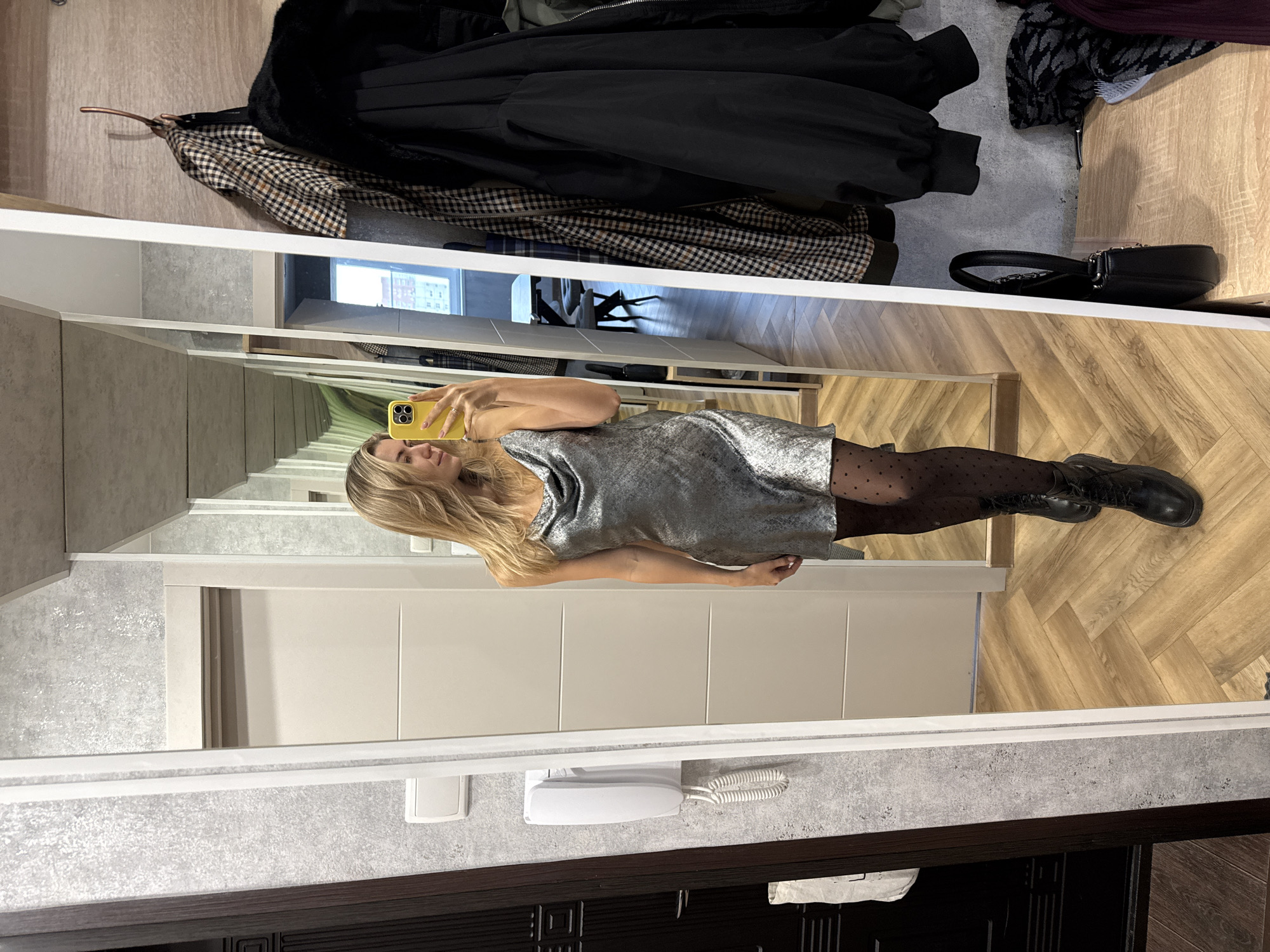 A full-length mirror captures a young woman taking a mirror selfie in what appears to be a modern entryway or hallway. The woman, with long blonde hair, is holding a bright yellow smartphone in her right hand, positioned to capture her reflection. She is wearing a short, shimmering silver or light grey dress, black sheer tights with small polka dots, and black lace-up boots. Her left arm is slightly bent, and her left hand is resting near her hip.
In the upper part of the image, above the mirror's reflection, several items of clothing are hanging on hooks against a wooden panel. These include a black coat or jacket and a plaid (brown, beige, black) jacket. To the right of the hanging clothes, a small black handbag with a chain strap rests on a surface.
The "real" environment, visible in the foreground and background outside the mirror's reflection, features a light grey textured wall, a dark brown wooden trim at the bottom, and light-colored wooden flooring laid in a herringbone pattern. A white landline telephone with a coiled cord is mounted on the textured grey wall below the mirror. The reflected environment within the mirror shows light-colored walls, possibly doors, and the same herringbone wooden flooring. No discernible text is visible in the image. The scene suggests an indoor setting, likely in a home or apartment.