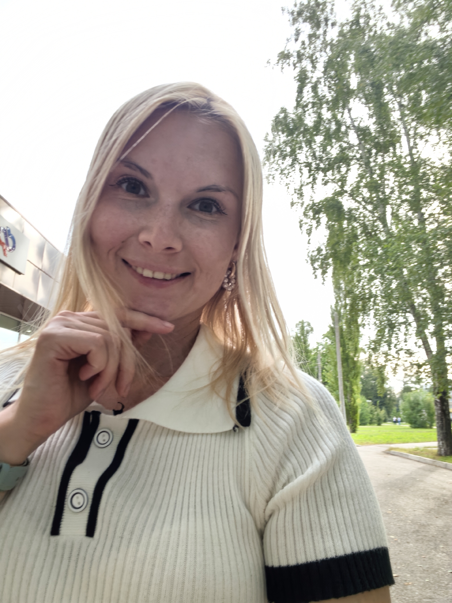 A woman with blonde hair and light skin, wearing a white ribbed shirt with black trim and buttons, is taking a selfie outdoors. She has a light-colored watch on her left wrist and wears pink earrings. Her right hand is on her chin, and she is smiling. Behind her, a building with a blue and red logo is visible on the left, and trees line a path on the right. The sky is bright and overcast. The location is likely Sterlitamak, Russia, based on the provided information.