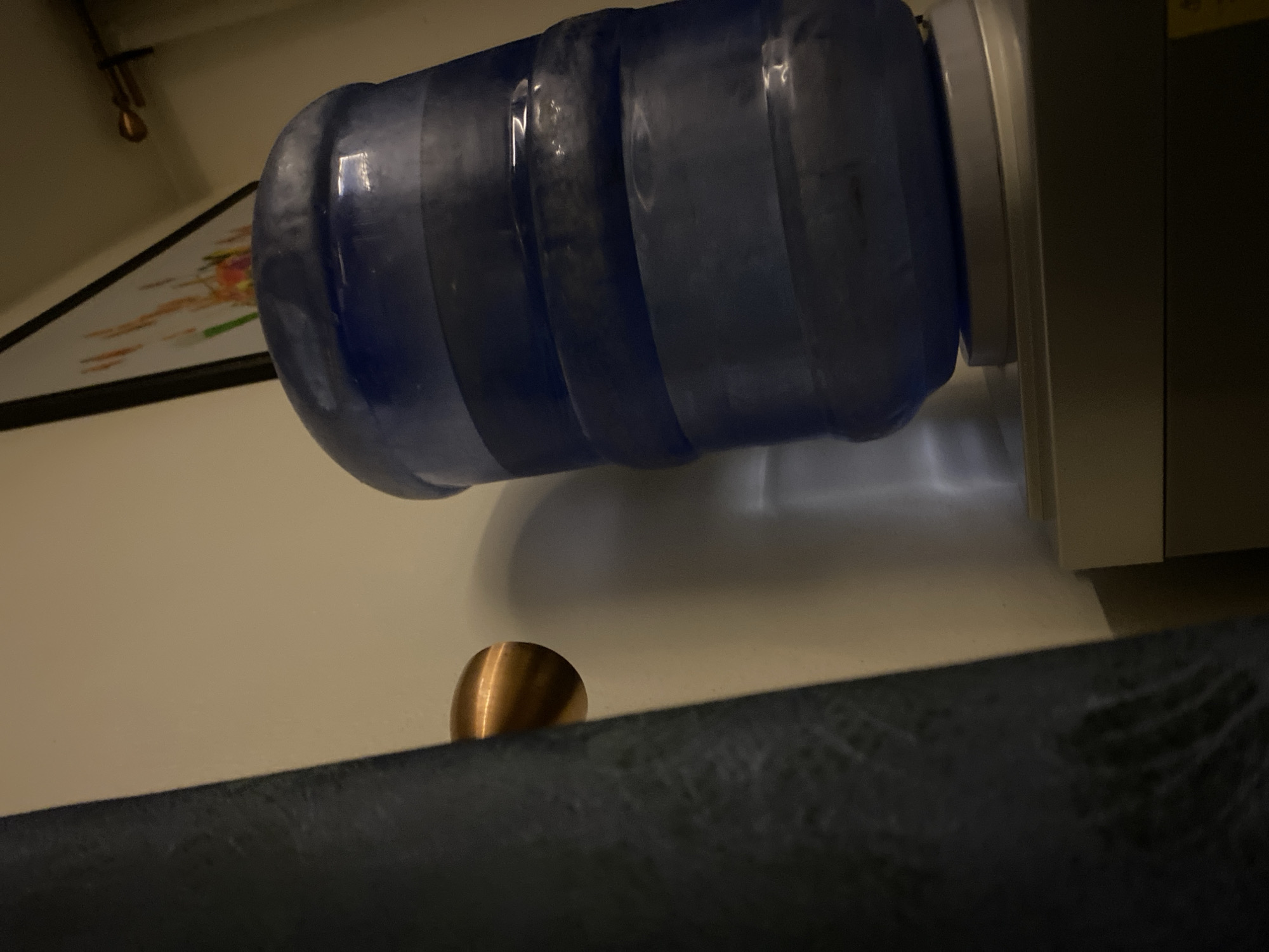 The image shows a water cooler with a large blue water bottle attached, placed on a light-colored surface. A small, copper-colored object, possibly a knob or handle, is partially visible near the edge of a dark, textured surface. The location, Abuja, Nigeria, and any details of people, actions, or interactions are not discernible from the image.