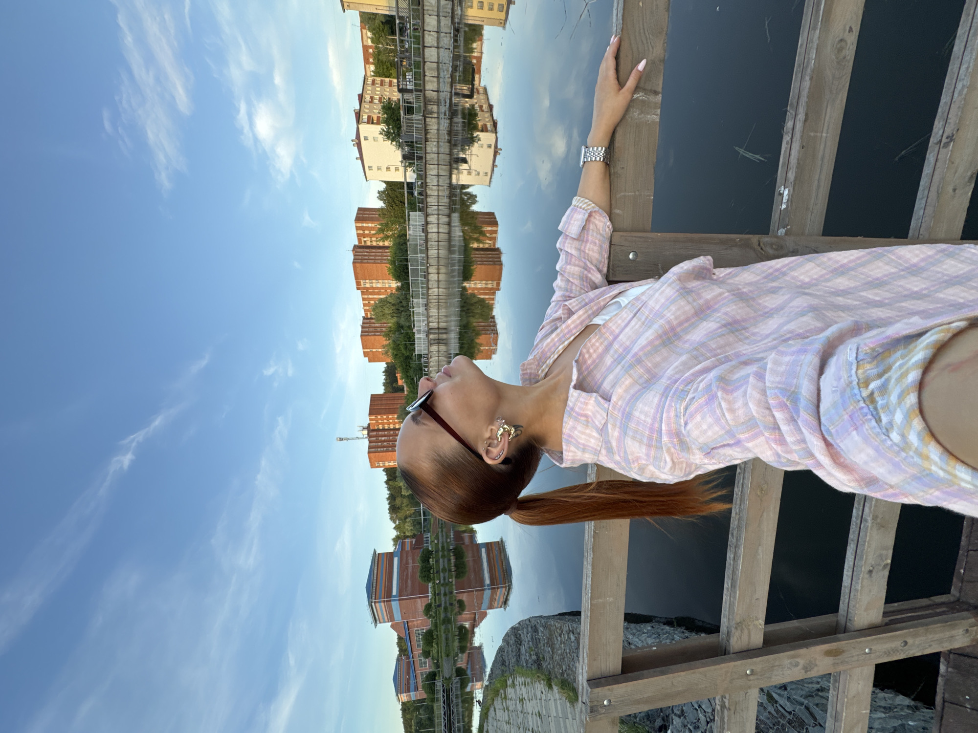 A woman with auburn hair in a ponytail, wearing a pink and white plaid shirt, sunglasses, and large earrings, leans on a wooden railing. She is looking up at the sky. The background shows a body of water reflecting buildings and the sky. The buildings appear to be apartment complexes and possibly an industrial building. The location is the city of Votkinsk, Russia. The sky is blue with wispy clouds.