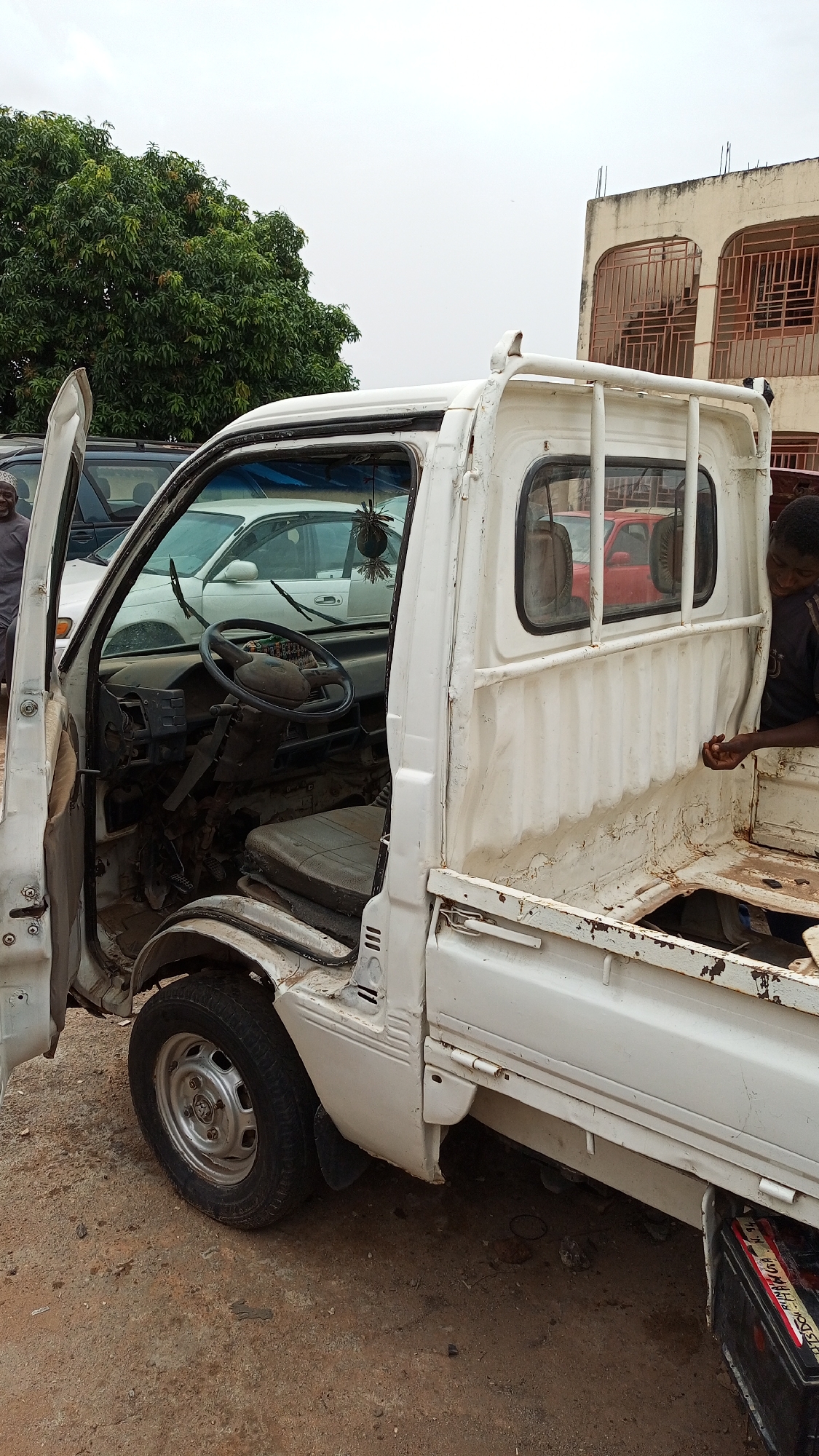 The image shows a dilapidated white pickup truck parked on a dirt lot. The driver's side door is open, revealing a worn interior with a grimy dashboard and seat. A small ornamental plant hangs from the rearview mirror. In the background, there are other vehicles, including a white sedan and a dark SUV. A man is visible near the front of the truck, while another man is seen looking inside the truck bed. A two-story building is partially visible in the background to the right. The location is identified as Kawo, Nigeria.