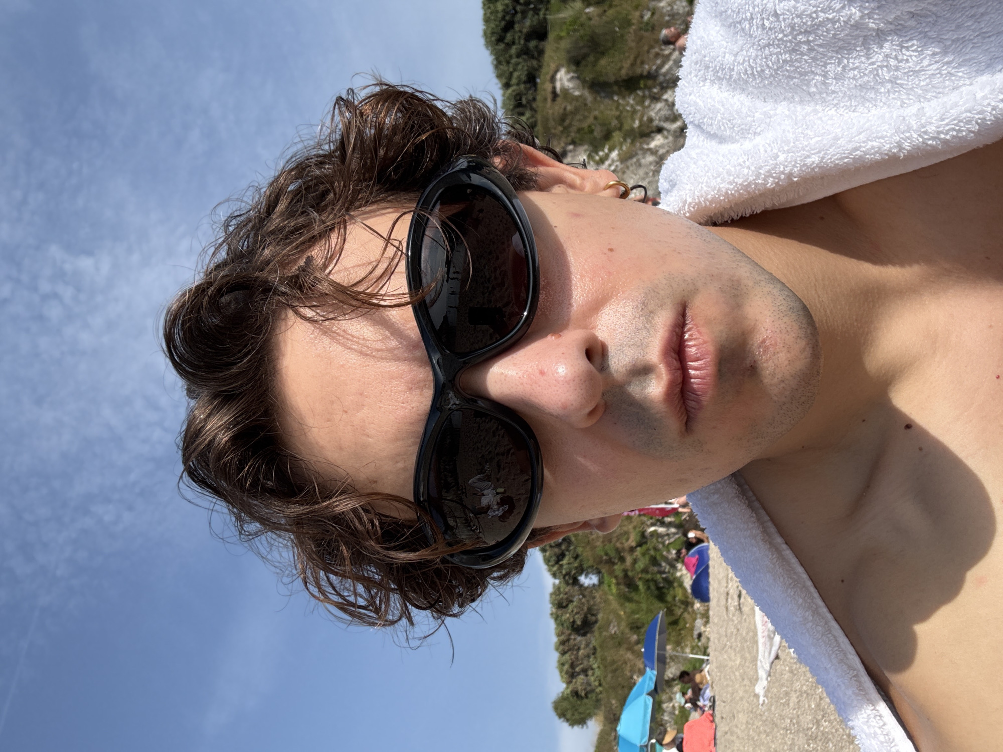 A young man is taking a selfie on a beach under a clear blue sky with sparse clouds. He is wearing dark, wrap-around sunglasses and a white towel draped over his shoulders. His medium-brown, wavy hair is windswept. The background shows a sandy beach with several beach umbrellas, some people, and a vegetated cliff face. The reflection in his sunglasses shows a person lying down on the sand. The image has a bright, sunny quality.