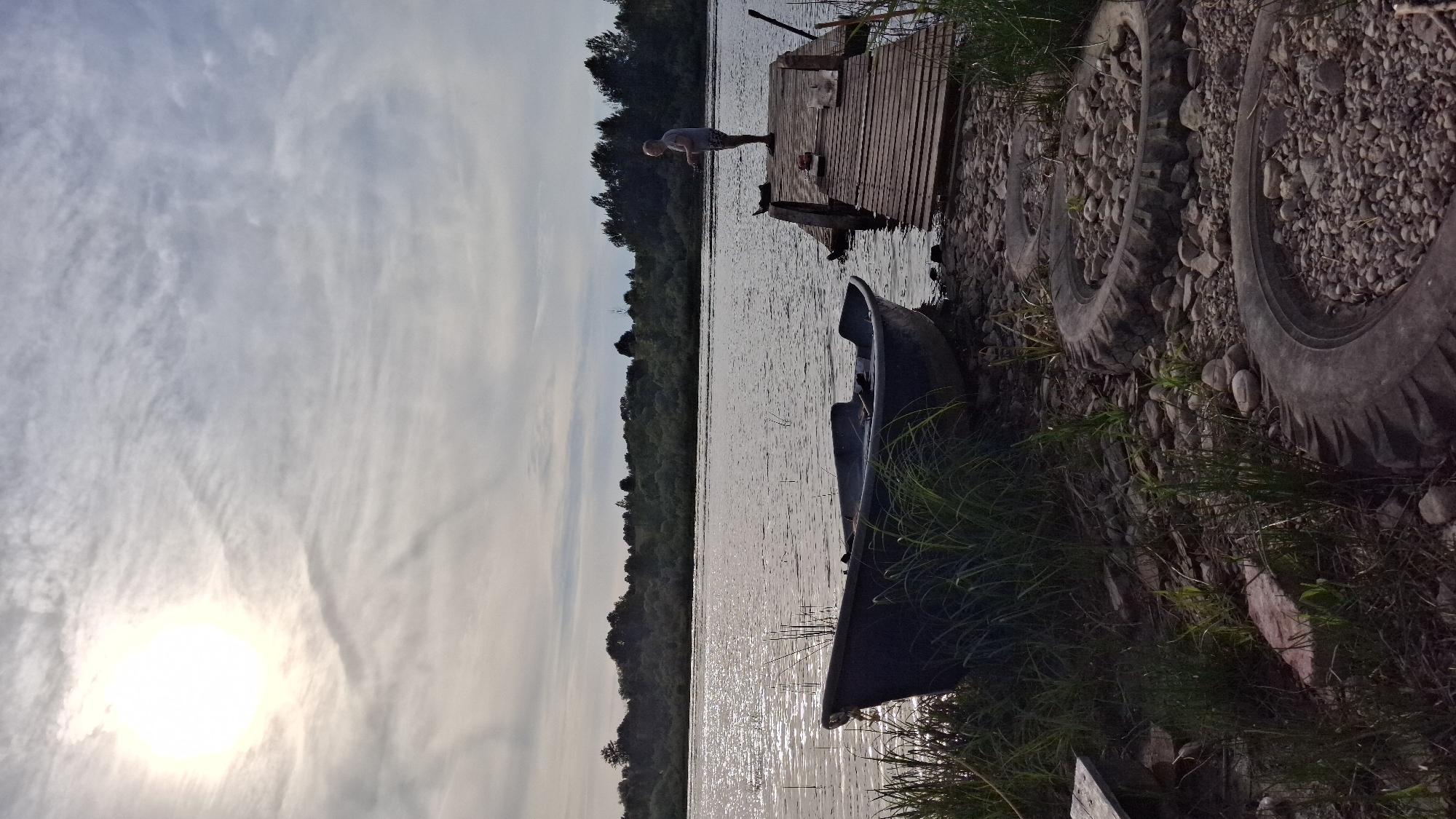 An elderly man is fishing from a wooden dock by a lake in Sitno, Russia. The sun is setting and casting a golden glow on the water. In the foreground, a small boat rests on the shore, with tall grass growing around it. Old tires filled with rocks are scattered on the ground, suggesting a rustic, lived-in area. The background is a dense forest that stretches across the horizon. The sky is cloudy, with the sun peeking through.