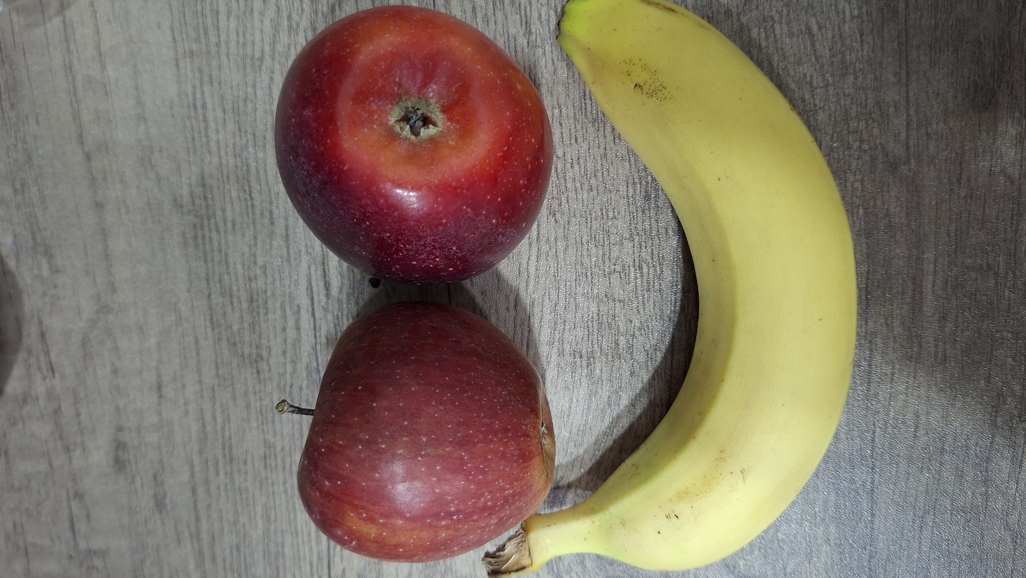 The image shows two red apples and a yellow banana on a wood-patterned surface. The larger apple is at the top, the smaller apple is below it, and the banana curves along the right side of the frame. The location is not deducible from the image.