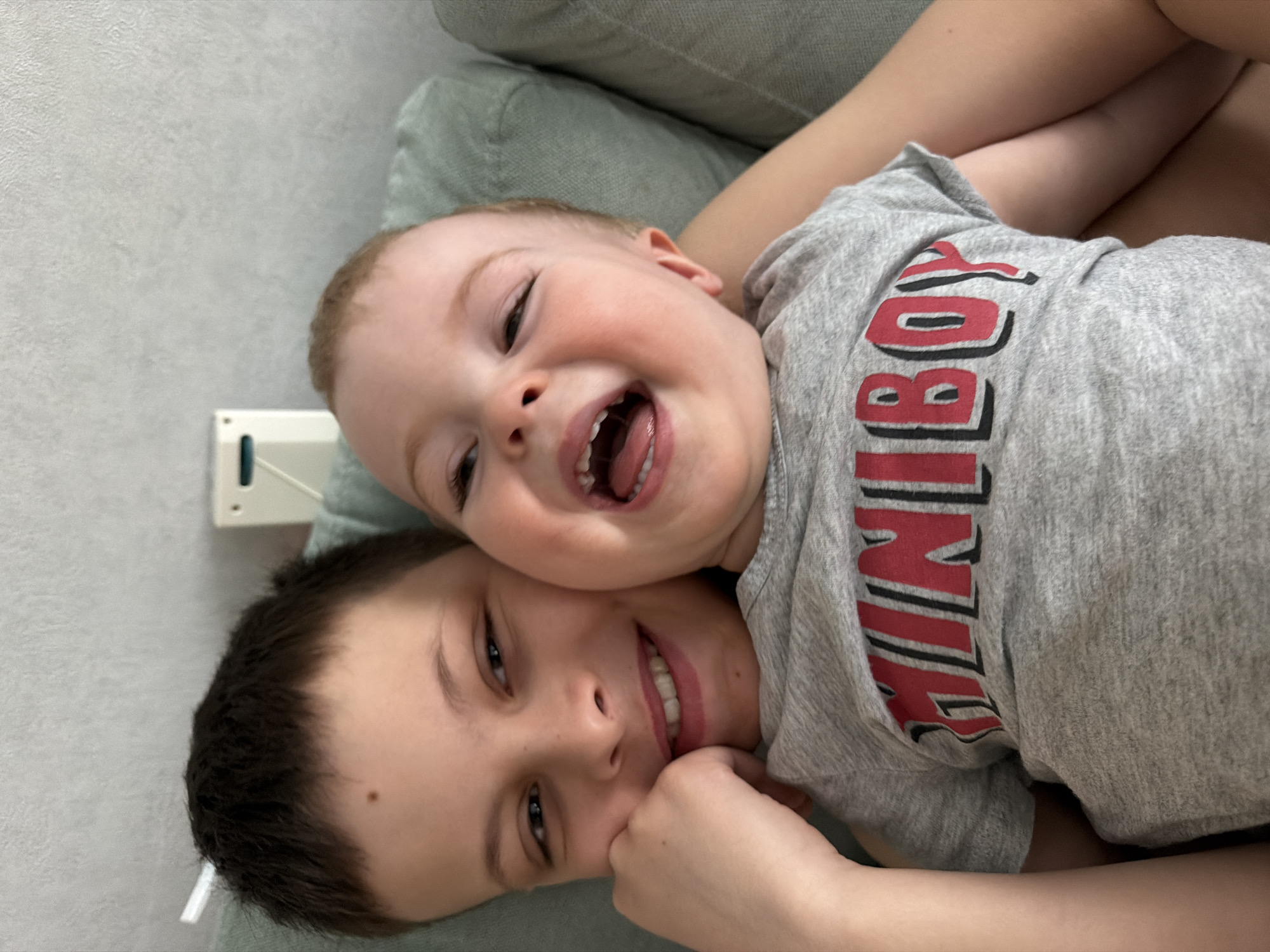 Two young boys are lying together on a soft, green-colored surface, likely a couch or cushion. The older boy, with dark hair and a mole on his cheek, is on the bottom, smiling and looking directly at the camera. The younger boy is on top of the older boy. He has light hair and is laughing with his mouth open, showing his teeth. He is wearing a gray t-shirt with red and black lettering that reads "MINIBOY". There is a white and green object on the wall in the background. The location is Votkinsk, Russia.