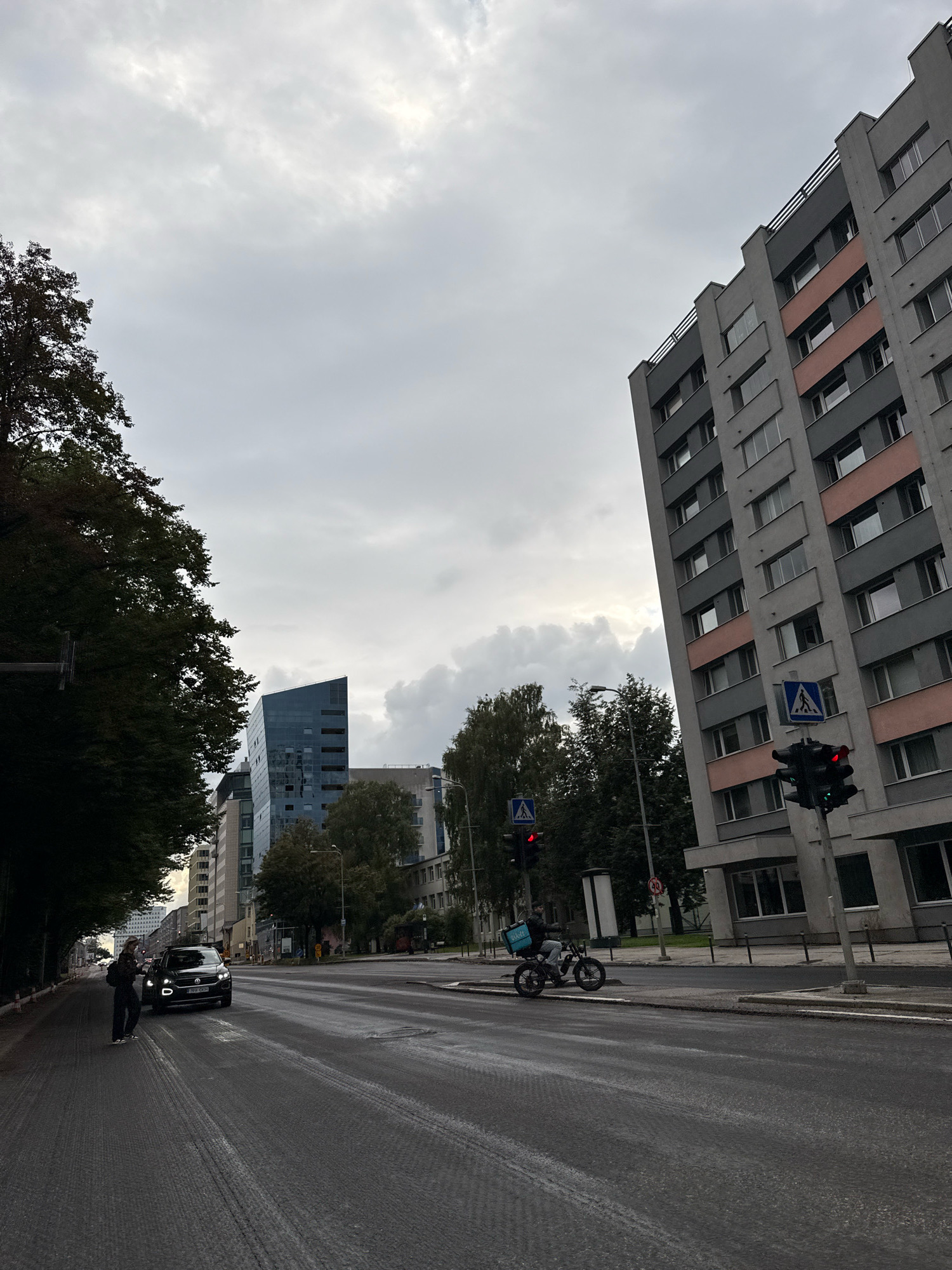 An overcast day in Tallinn, Estonia, with a street scene featuring modern architecture. On the right, a tall, grey building with pink accents rises into the sky, its facade marked by numerous windows. A traffic light on a pole, displaying red and green signals, stands in front of it. Nearby, a blue pedestrian crossing sign is visible. Further down the street, a person on a dark-colored electric scooter with a bright blue "Bolt Food" delivery bag is moving away from the camera. On the left side of the street, a dark-colored car is parked. A person with a backpack is standing on the sidewalk near the car. Tall trees with dark green foliage line the left side of the street, creating a shadowy effect. In the background, a uniquely shaped, modern glass building with a blue tint stands out, reflecting the sky. The street is wet, suggesting recent rain, and the sky is filled with diffused light from the clouds.
