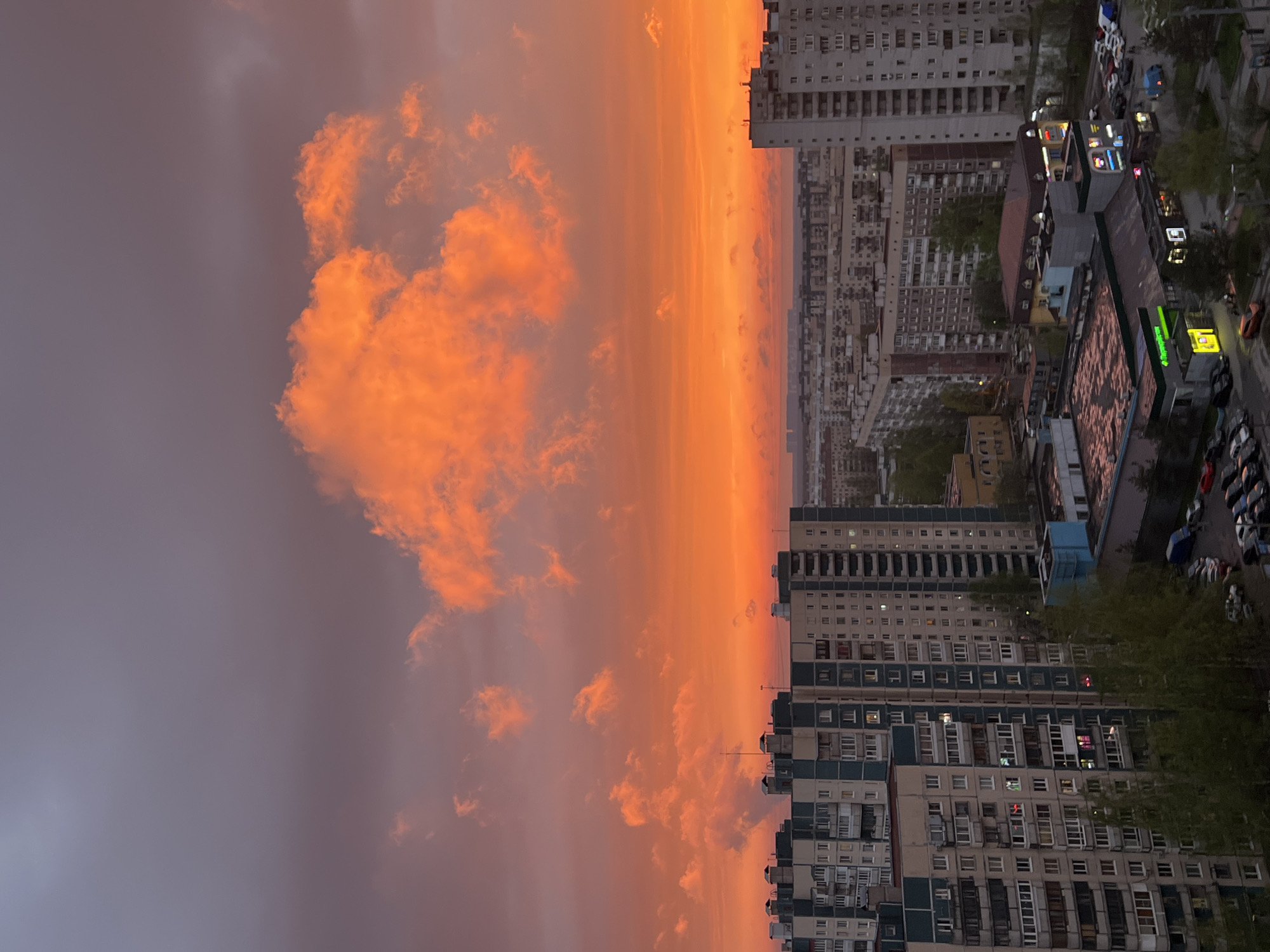 The image presents an elevated cityscape at dusk or dawn. The focal point is the sky, displaying an intense orange and red gradient near the horizon, transitioning to darker grey clouds overhead. Adjacent to the right is a high-density urban environment with numerous apartment buildings of varying heights and architectural styles, indicative of Soviet-era construction. Street-level features include parked cars, trees, and commercial structures with visible signage. Specific details visible through the windows of buildings and signage suggest the location is Saint Petersburg, Russia, due to the style of architecture.