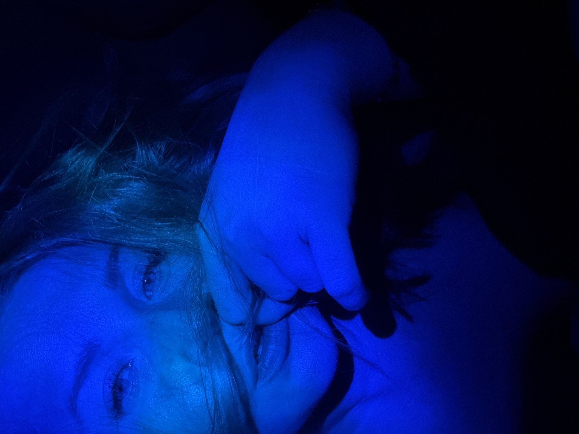 The image presents a close-up, low-light portrait of an individual, likely female, illuminated predominantly by a vivid blue light source. The subject's face occupies the upper-center to mid-left of the frame, with her eyes directly engaging the viewer. Her pupils appear dilated, and irises, while blue-tinted by the light, seem light-colored. Defined eyebrows are visible above.
Strands of light-colored hair, appearing blue under the illumination, fall across her face, partially obscuring the nose and mouth area. A left hand, also saturated in blue light, is positioned to the lower-left, with fingers gently curled. The index finger and thumb are seen pulling or holding strands of hair near her partially open mouth. The mouth is slightly agape, revealing a glimpse of teeth. The lower lip is prominent. A small dark mark or blemish is visible on the back of the hand near the knuckles.
The subject's upper chest and shoulder are faintly visible on the lower-left, also illuminated by the blue light. The background is entirely dark and undifferentiated, providing no contextual information. The location of the photograph cannot be determined from the visual evidence provided; there are no discernible features to indicate it is Veliky Novgorod, Russia, or any other specific geographical area. The overall ambiance is intense and dramatic due to the monochromatic blue lighting and tight cropping.