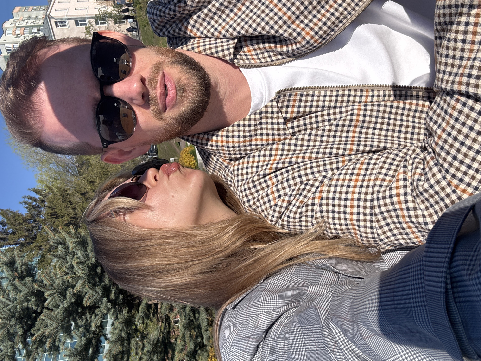 A selfie captured outdoors shows a man and a woman in close proximity. The man, positioned at the top of the frame, has fair skin, a trimmed beard, and sunglasses. He wears a white t-shirt beneath a brown and white checkered jacket. The woman, located below him, also wears sunglasses. She has light brown hair and a grey and white checkered jacket. They appear to be in a park or green space, with trees visible in the background. In the far distance, indistinct buildings can be seen. The overall scene suggests a casual moment between the two individuals in an urban environment.