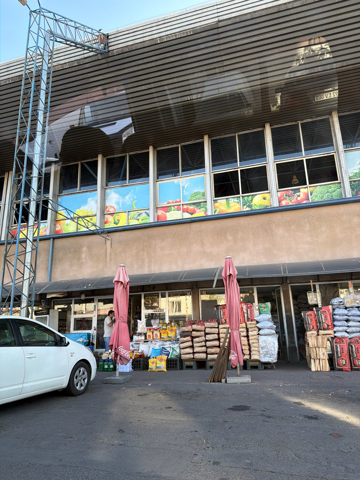 The image depicts an outdoor market scene in Yerevan, Armenia, likely during daylight hours. In the foreground, a white car is parked on an asphalt surface. To the right of the car, a market stall is set up with various goods displayed outside. These include stacks of bagged charcoal or similar fuel, wooden logs, and smaller bags of produce or other items. A large pink umbrella stands near the stall.
Behind the market stall, the facade of a building is visible. It features a series of large windows displaying colorful graphics of fruits and vegetables against a sky backdrop. The building has a covered awning extending over the market area. A person, appearing to be a vendor, is standing inside the market stall area.
To the left of the market stall, a tall metal structure, possibly a lamppost or part of a larger infrastructure, extends upwards. The overall environment suggests a commercial area in a city. The presence of Armenian script on some of the bagged goods confirms the location as Yerevan.