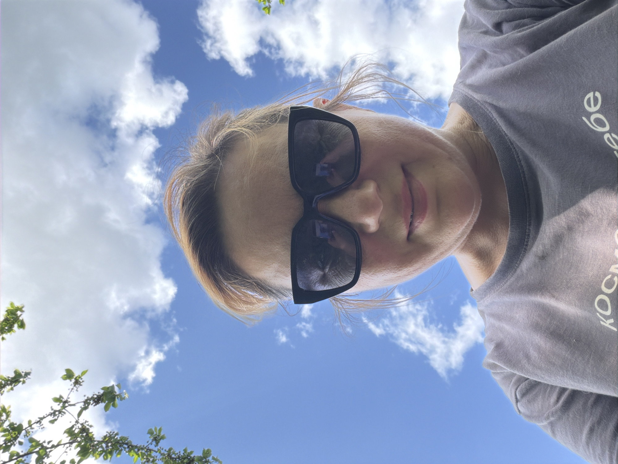 A woman is looking down at the camera, wearing sunglasses and a grey t-shirt with the word "КОСМОСЕ" printed on the sleeve. The background shows a bright blue sky with white clouds and some green foliage in the lower left corner. Based on the text on the shirt, the woman is possibly located in Leningradskaya, Russia.