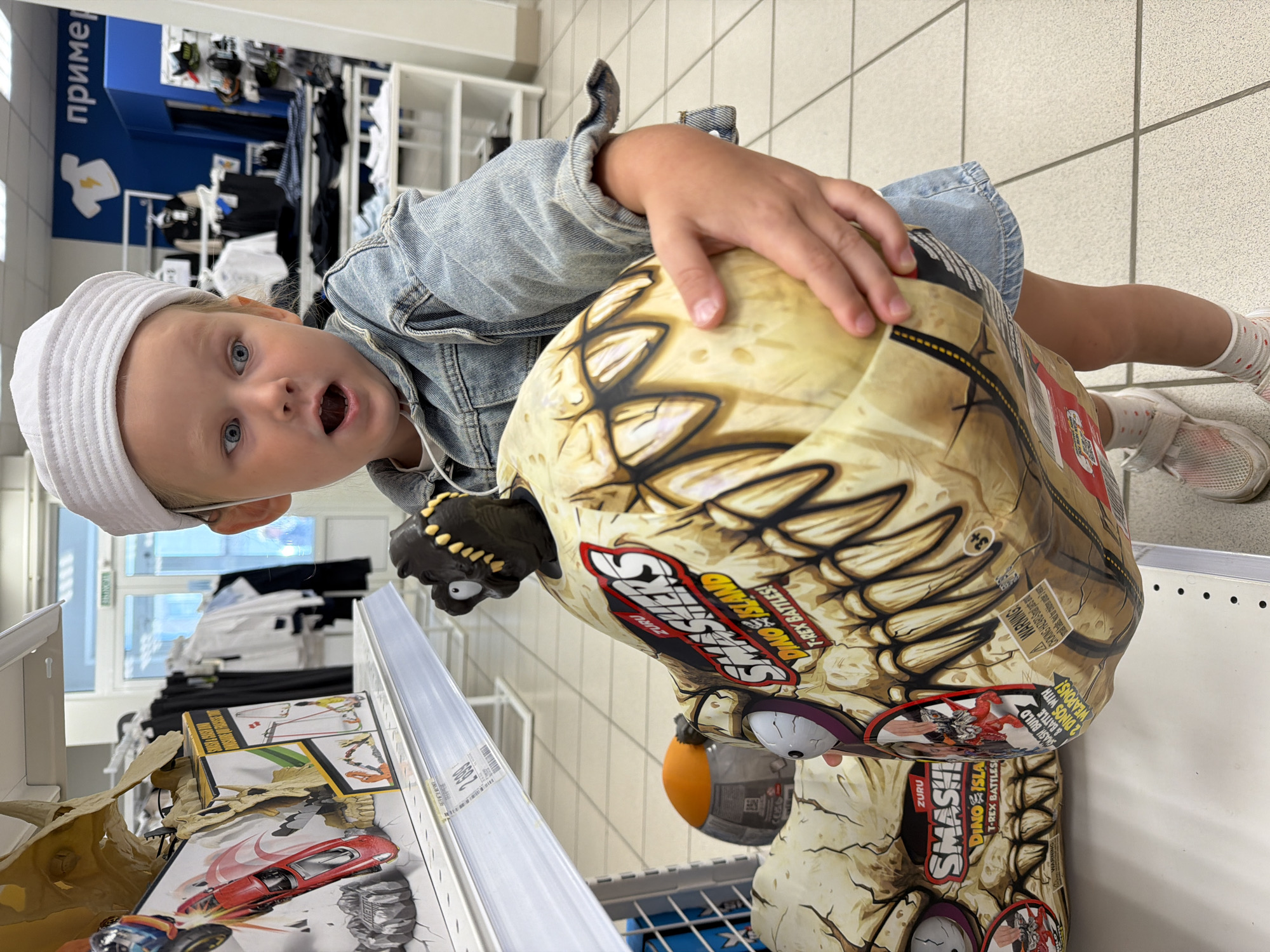 A young child, wearing a white sailor hat and a denim jacket, is standing in a store, holding a large, spherical toy with a dinosaur-themed design. The toy is light brown with images of dinosaur teeth and the brand "ZURU SMASHERS DINO ISLAND T-REX BATTLES". The child has a surprised expression on their face, with wide eyes and an open mouth. In the background, shelves are stocked with various items, including clothing and other toys. A red toy car is visible on a shelf to the left. The floor is tiled. The store appears to be part of a chain, with signage in Russian that includes the word "примерка" (fitting room). Based on the store's aesthetic and the Russian language present, this scene is likely located in Kamyshin, Russia.