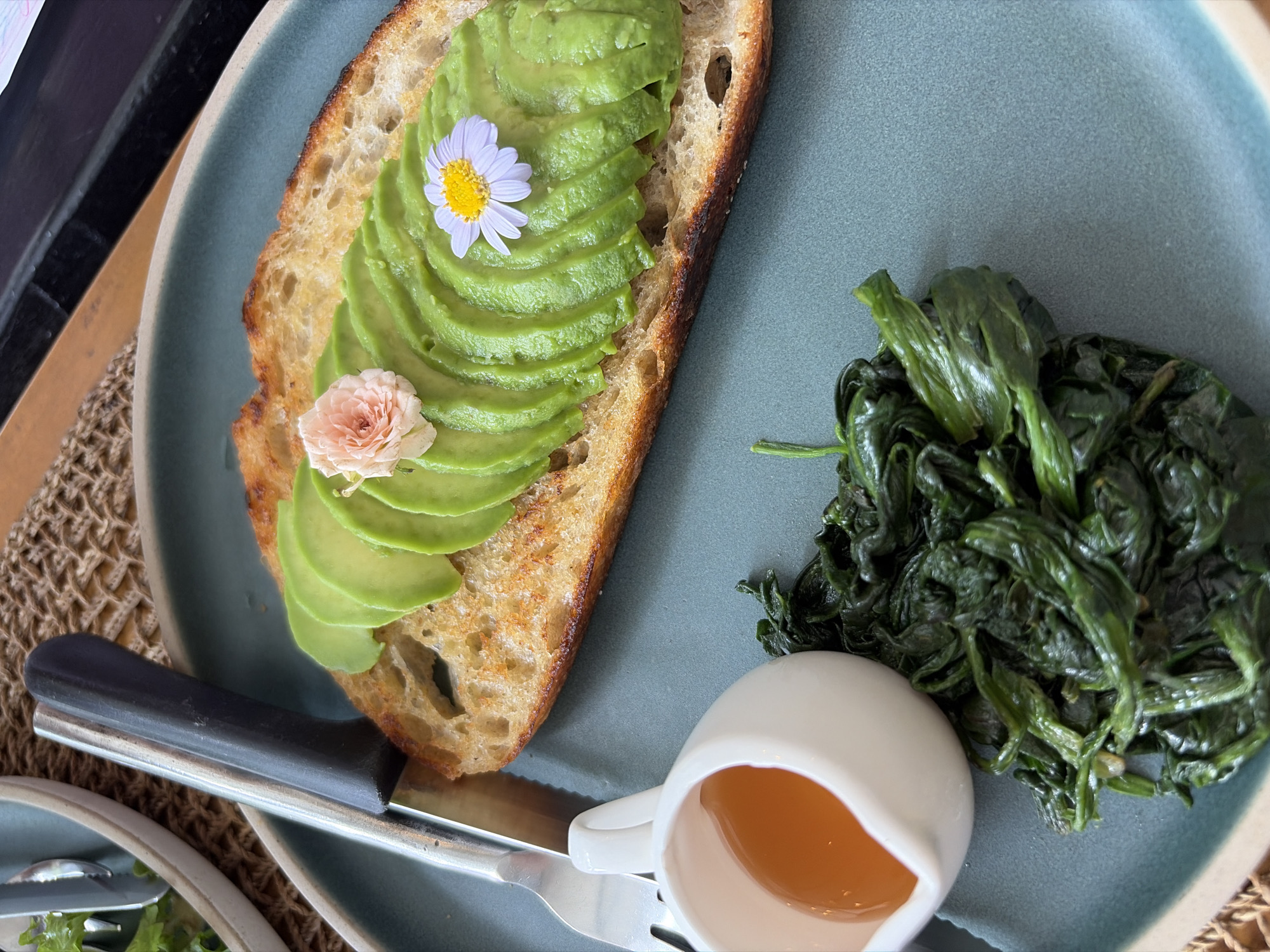 A close-up, overhead shot shows a plate of food on a table in Baan Bo Phut, Thailand. The plate contains a slice of toasted sourdough bread topped with fanned-out avocado slices, a small pink flower, and a single white daisy. To the left of the toast is a small white ceramic pitcher filled with amber-colored liquid, likely honey or syrup. Below the pitcher and to the right is a serving of cooked spinach. A fork and knife are partially visible on the left side of the frame. The table surface appears to be covered with a woven placemat and the plate rests on a larger teal-colored plate. The background is blurred, but suggests an indoor setting.