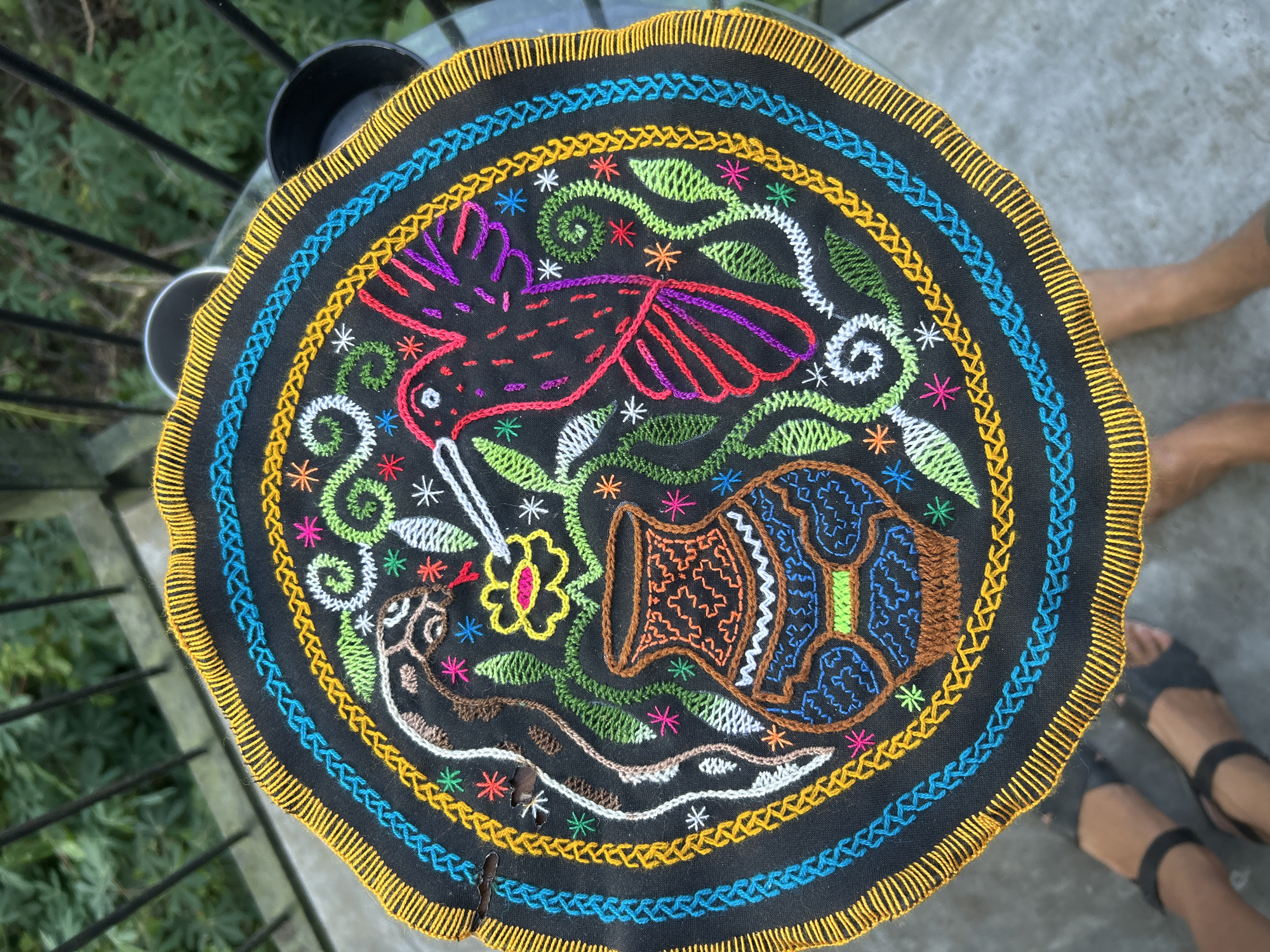 A vibrant, circular embroidered textile, likely a mat or decorative cover, is prominently displayed on a dark, glossy table, possibly made of glass. The embroidery, crafted on a black fabric background, features a rich palette of colorful threads.
The central design showcases a dynamic natural scene. A red-bodied bird, possibly a hummingbird, with striking purple and orange wings and a long beak, appears to hover above a yellow and pink flower, suggesting it is feeding. To the right of the flower, a brown and blue patterned pot or vessel is depicted, while a white and brown snake-like creature coils towards the bottom left. These main elements are enveloped by an assortment of green, white, and multi-colored floral and leafy motifs, including spirals and star-shaped accents, which fill the space with intricate detail.
The central artwork is framed by multiple concentric borders of varying stitch patterns in bright yellow and blue threads. The outer edge of the textile is finished with a distinct scalloped border in yellow stitching.
The setting appears to be outdoors during daylight hours. In the top left background, lush green foliage is visible through a dark metal railing, suggesting a patio, balcony, or garden area. To the bottom right, the bare legs and feet of a person, wearing dark sandals, are visible on a light gray concrete or tiled surface, indicating their presence near the table. The image exudes a sense of traditional artistry in a casual, outdoor environment, consistent with the provided location context of Baan Wok Tum, Thailand. No text is visible in the media.