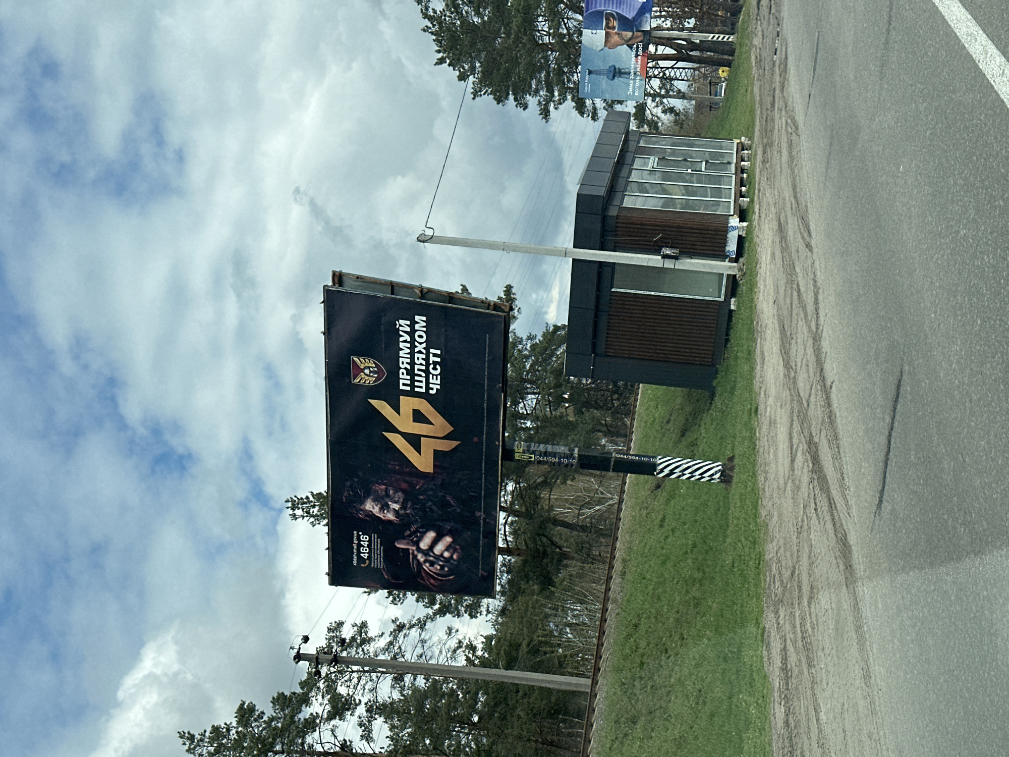 The image shows a roadside view in Hostomel, Ukraine, featuring a billboard advertising recruitment for the Ukrainian Armed Forces. The billboard displays the number "46" prominently, along with the text "ПРЯМУЙ ШЛЯХОМ ЧЕСТІ" (Follow the Path of Honor), a depiction of a soldier, and a contact number. A modern bus stop shelter is situated next to the billboard along the road. In the background, there are power lines, trees, and another billboard advertising the armed forces. The sky is overcast. Tire tracks are visible on the grassy shoulder of the road.