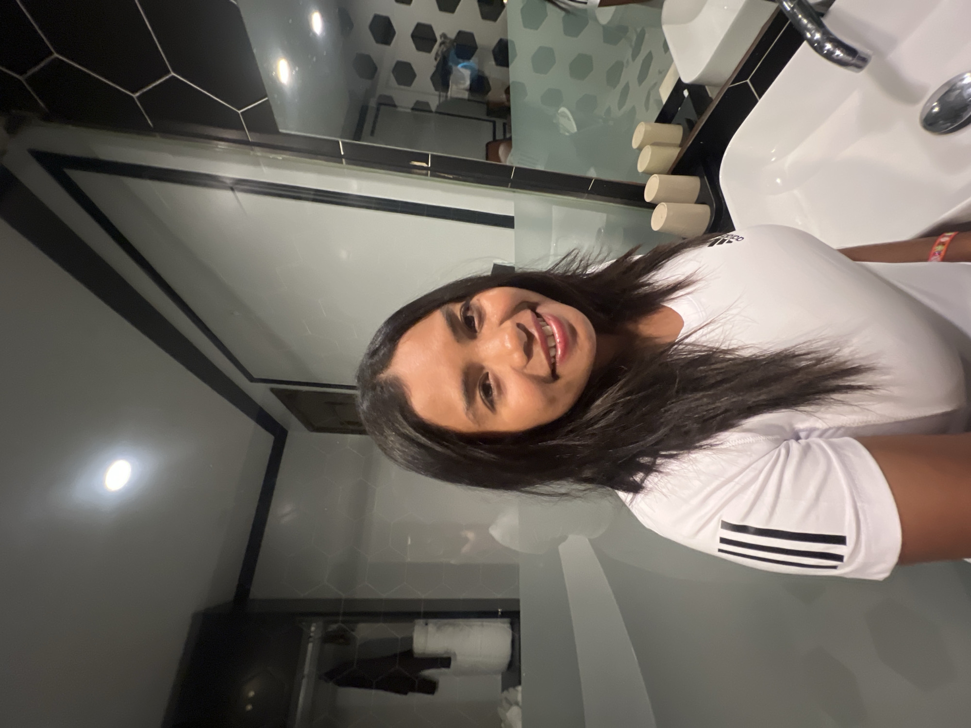 A woman with long, dark hair smiles at the camera in a modern bathroom. She is wearing a white t-shirt with black stripes on the sleeve and an Adidas logo on the chest. In the background, a large mirror reflects the bathroom, revealing hexagonal patterned tiles on the wall and a sink. The overall impression is one of a stylish and well-lit space. There is no information available about the specific location in Hanoi, Vietnam, or any interactions between people.