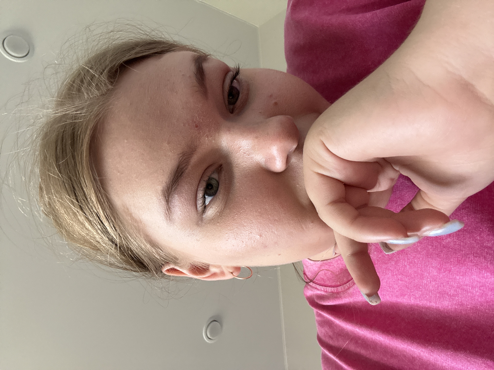 A close-up, angled view depicts a woman with light blonde hair and fair skin. She wears a pink shirt, and her hand is raised near her face. She has green eyes and wears hoop earrings. The background is a plain, light-colored wall with two circular objects, likely light fixtures. The location is not discernible from the provided image. The scene does not depict any specific actions or interactions.