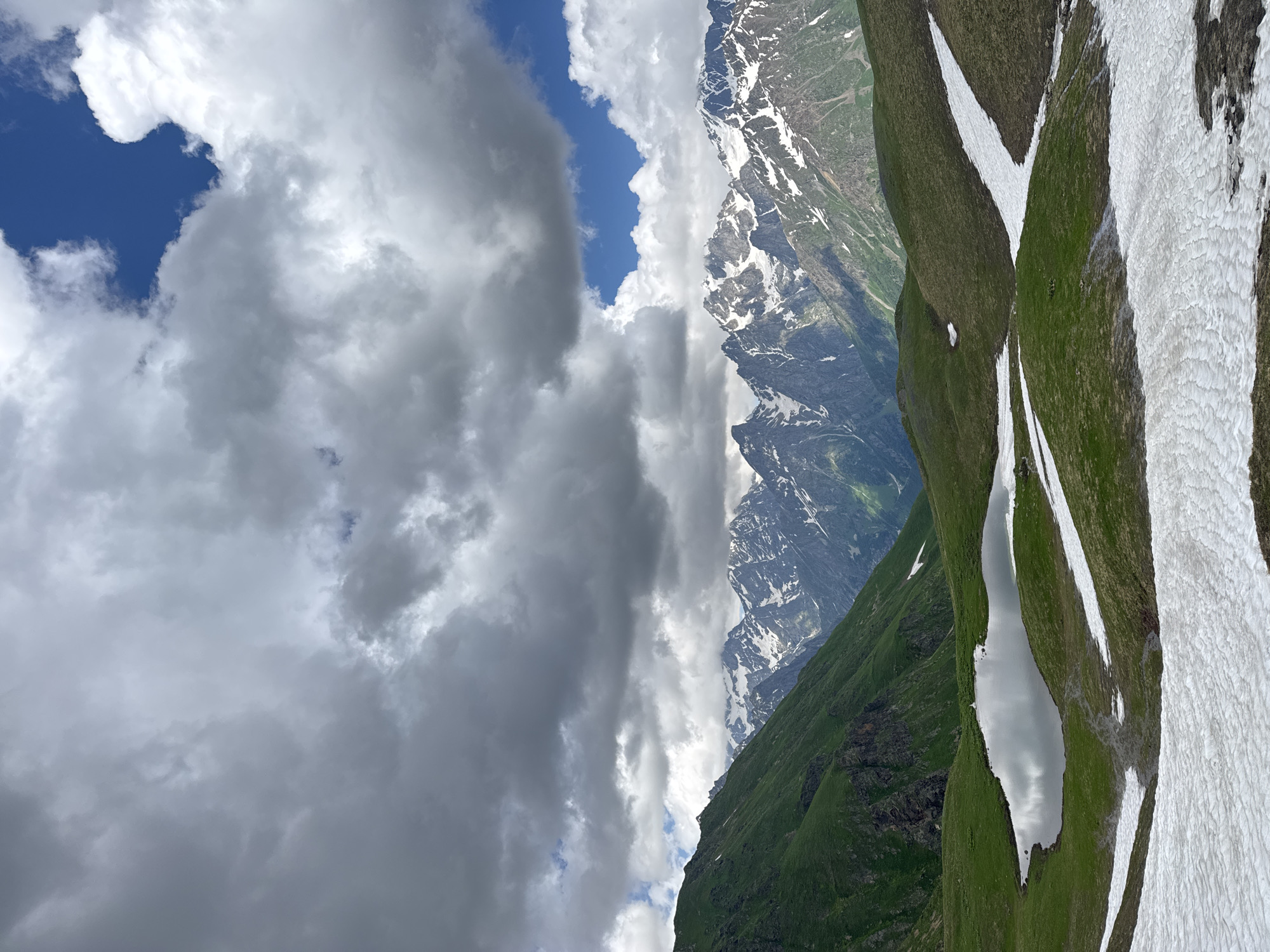 An aerial view of the Chalaadi Glacier in Mestia, Georgia, showcases a dramatic mountain landscape. The sky above is a canvas of dramatic clouds, with patches of bright blue sky peeking through. In the foreground, rolling green hills are dotted with patches of melting snow. A small, still lake reflects the cloudy sky. In the mid-ground, a range of majestic, snow-capped mountains rise, their jagged peaks piercing the sky. The slopes of the mountains are covered in lush green vegetation. The scene is serene and wild, conveying the raw beauty of the Georgian Caucasus.