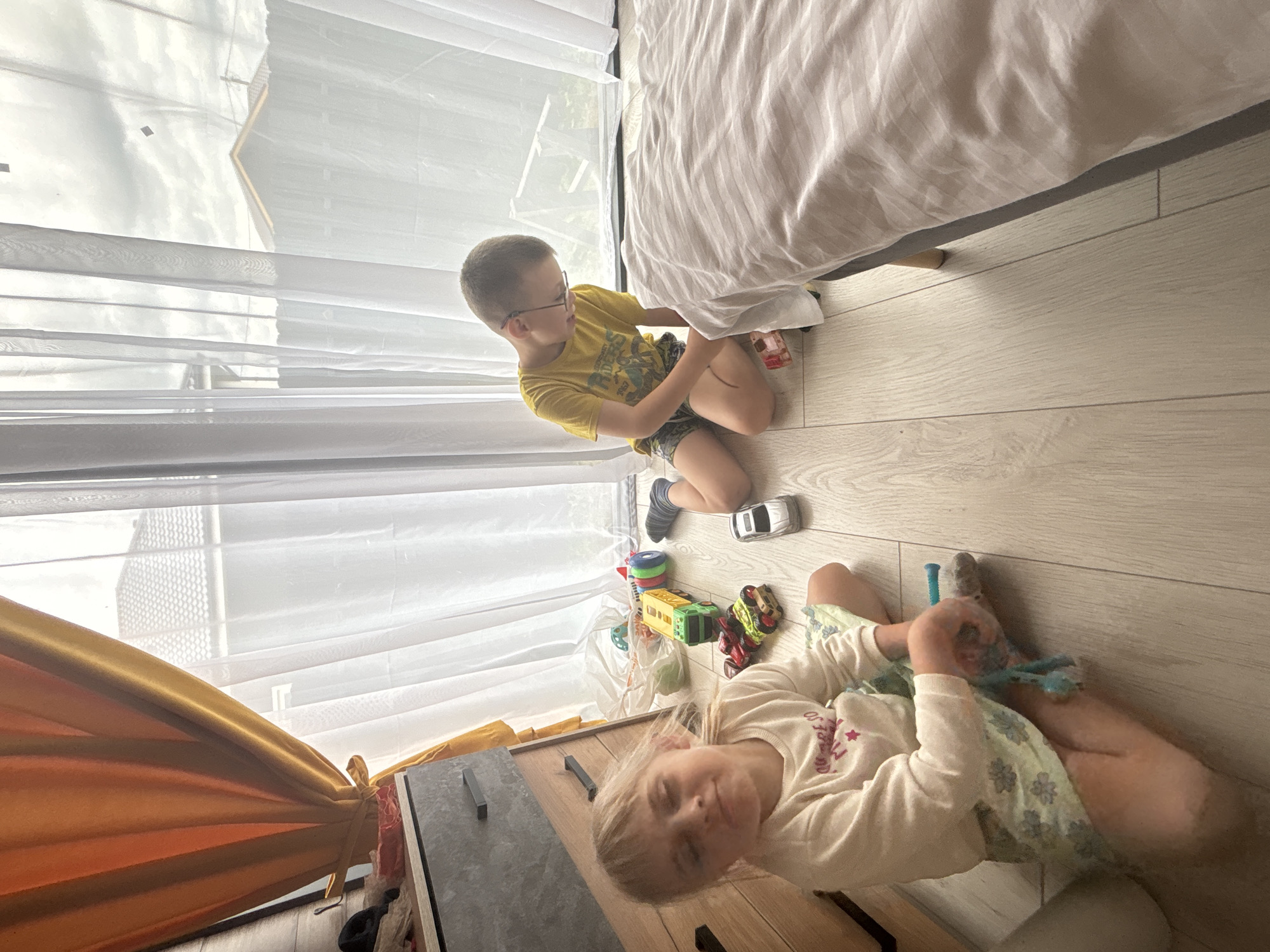 Two children are on the floor in a room with light-colored wood flooring. A young girl, appearing to be around 4-6 years old, sits cross-legged in the foreground on the left, facing the camera with her eyes closed and a slight smile. She is wearing a long-sleeved white shirt with pink lettering and a light green skirt with a floral pattern. She holds a blue toy in her hands. To her right, a young boy, likely 7-9 years old, kneels on the floor, facing away from the camera and towards a bed. He is wearing a yellow t-shirt with a graphic print and patterned shorts. He is wearing glasses and appears to be drawing on his leg with a marker or similar object. Scattered on the floor between the children are several toys, including a yellow and green toy truck, a red and yellow toy vehicle, a silver toy car, and other colorful plastic toys. A large bed with white striped bedding is positioned to the right of the frame. In the background, sheer white curtains are drawn across a window, revealing glimpses of a residential exterior with a house roof and fence. The room appears to be a bedroom or playroom. The lighting suggests it is daytime, with natural light coming through the window.