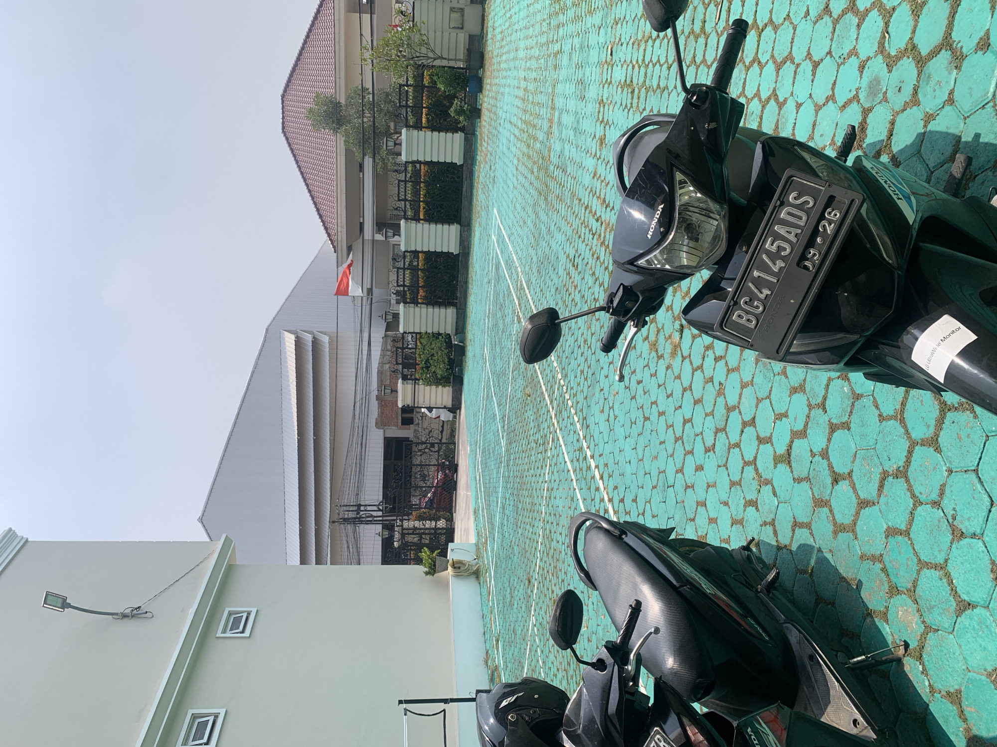 The image captures an outdoor scene, possibly in Palembang, Indonesia, given the license plate format. In the foreground, two black scooters are parked on a teal-colored hexagonal paver surface that appears to be marked as a badminton court. The scooter on the right is closer to the camera and displays a license plate with the text "BG4145ADS 09.26". Its front is angled towards the viewer, showing the headlight and handlebars. The scooter on the left is partially visible, with its front wheel and a black helmet resting on its seat.
In the midground, a light-colored building with white trim and small square windows is on the left. Beyond the paver court, a fence with decorative pillars and gates encloses a property with a red-tiled roof house and some greenery. A large, industrial-style building with a sloped roof is visible behind the fence. A red and white flag, likely the Indonesian flag, is prominently displayed on a flagpole near the roofline of the house. The sky is bright and overcast, suggesting daytime. The overall impression is one of a functional outdoor space, possibly a residential compound or a facility with recreational areas.