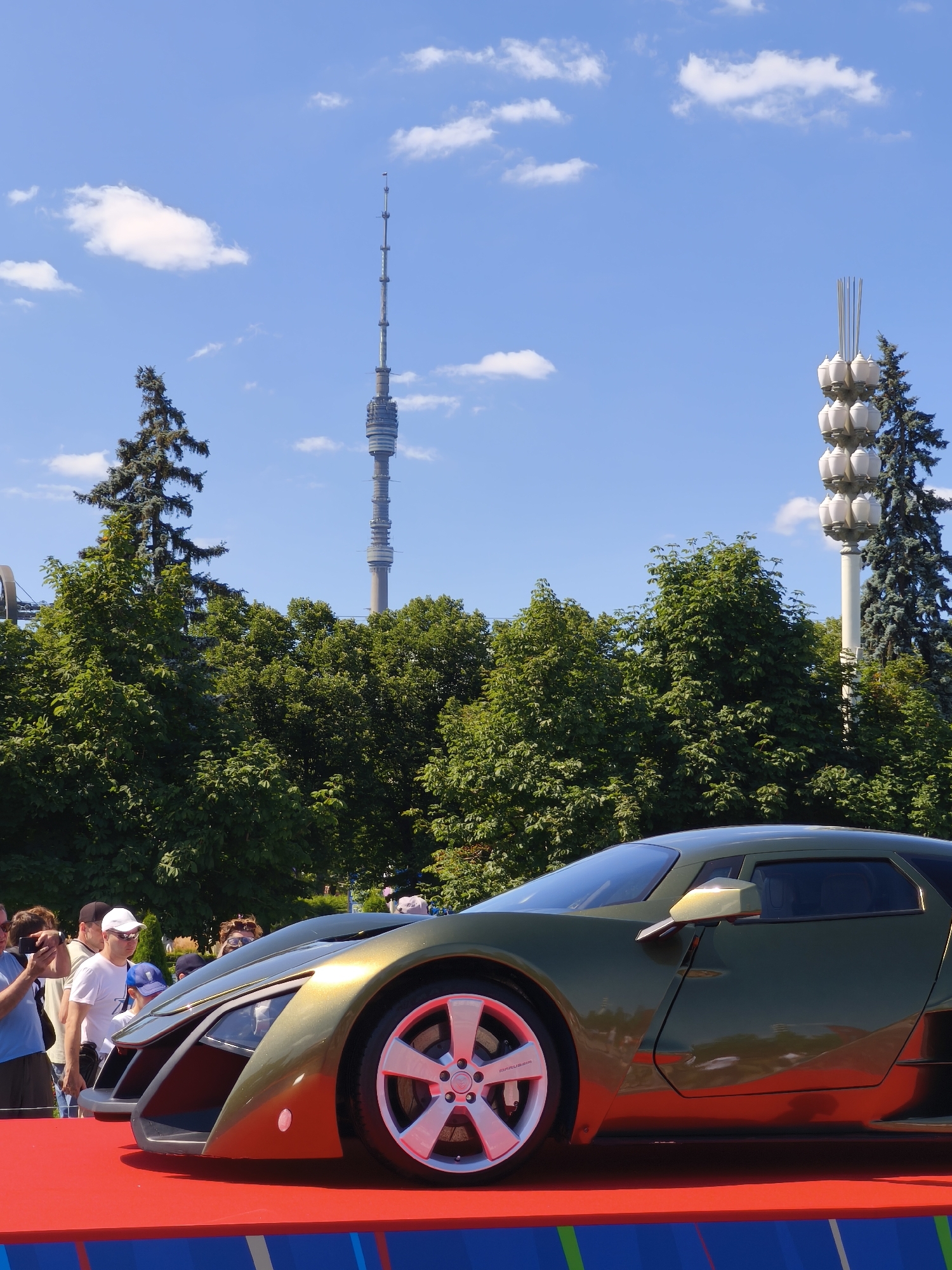 An outdoor, eye-level, medium shot features a futuristic, olive-green concept car displayed on a red platform. The car's sharp, angular design and prominent silver wheel are in focus. Behind the car, a crowd of people observe the display. The background reveals lush green trees, a tall, slender television tower in the distance, and a cluster of outdoor lamps against a bright blue sky with scattered white clouds. The television tower is identifiable as the Ostankino Tower, and the lamps are likely part of the Exhibition of Achievements of National Economy (VDNKh) complex, indicating the scene is located in Moscow, Russia.