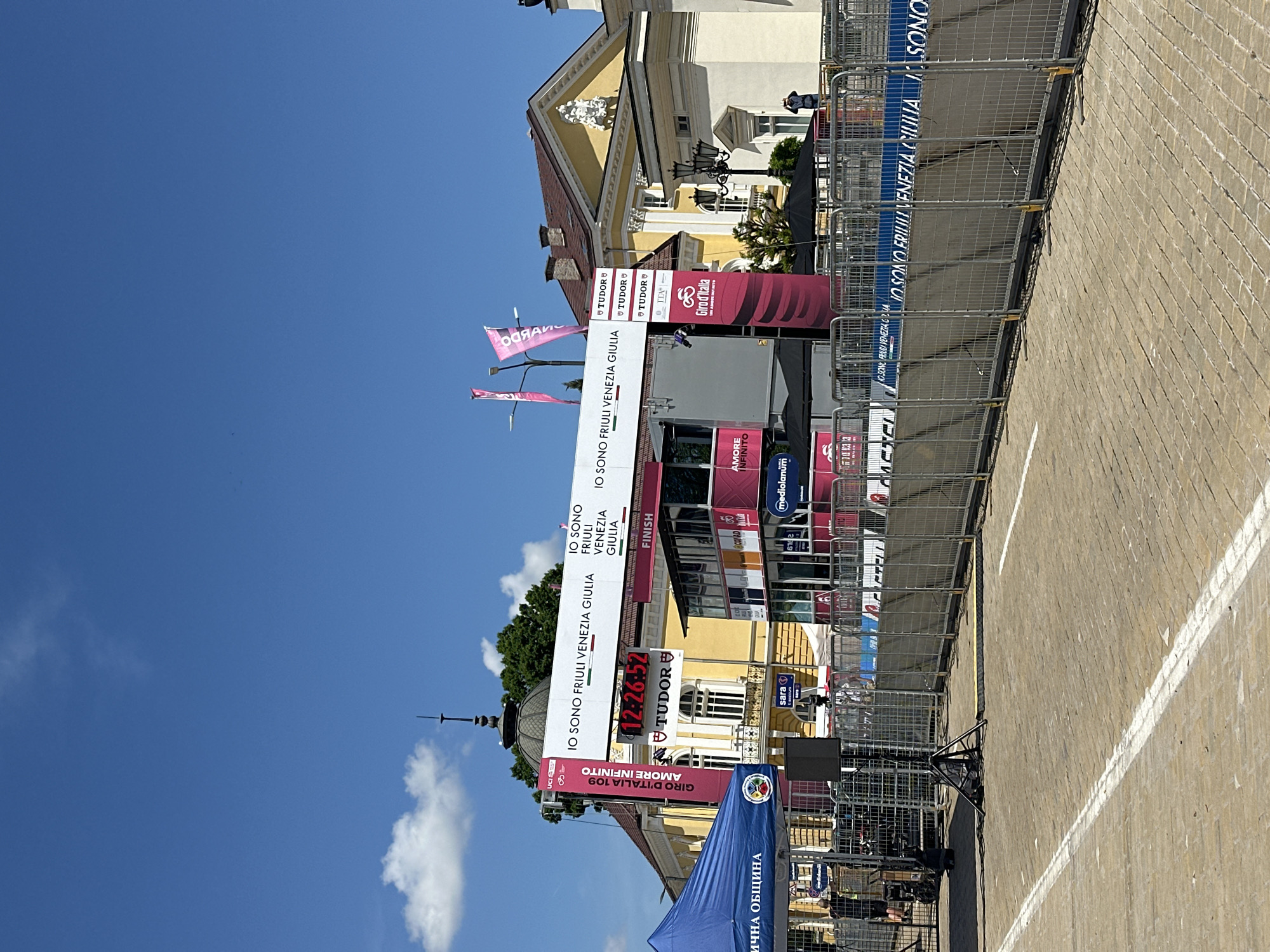 The image depicts the finish line area of a Giro d'Italia cycling race in Sofia, Bulgaria. Prominent features include barriers lining the race course, the finish line structure with banners displaying "IO SONO FRIULI VENEZIA GIULIA", "TUDOR", "Giro d'Italia", and "AMORE INFINITO". Logos of sponsors like "Mediolanum" and "Sara" are visible. Flags and tents are present in the surrounding area. The building behind the finish line area appears to be a historical structure. The sky is blue with some clouds. The road surface is paved. The displayed time is "12:26:52".
