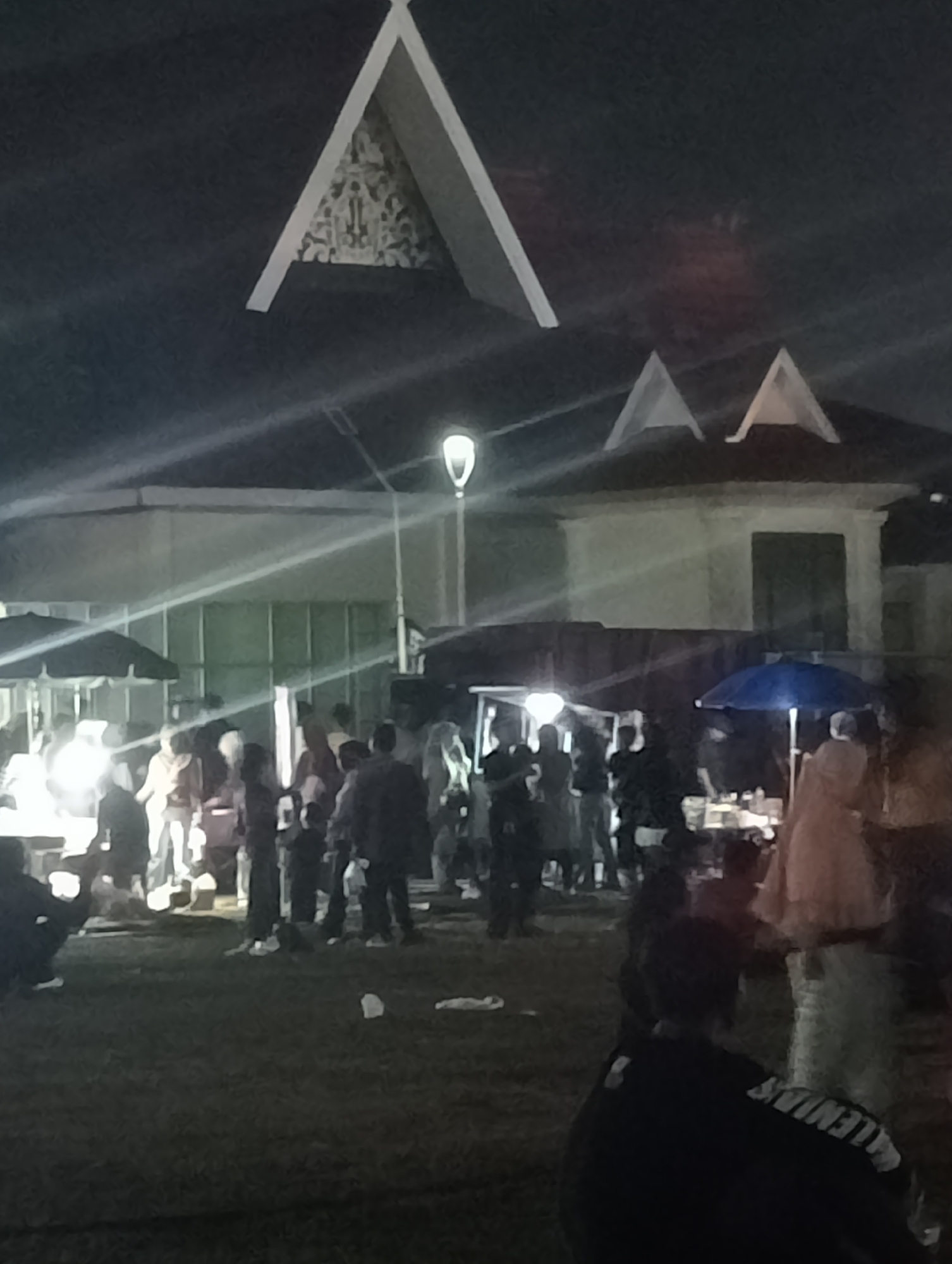 The image depicts a nighttime scene in Jambi City, Indonesia, likely at a public gathering or event. In the foreground, several individuals are visible, with one person in the lower right wearing a dark shirt with white lettering on the back. The middle ground shows a crowd of people mingling, some standing under umbrellas, and what appears to be food stalls or vendor tents. Bright lights illuminate some areas, casting long shadows. In the background, a building with a distinctive triangular roof design is prominent, and another building with a similar roof structure is also visible. Streetlights are present, contributing to the overall lighting of the scene. The ground appears to be covered in grass, with some scattered debris.