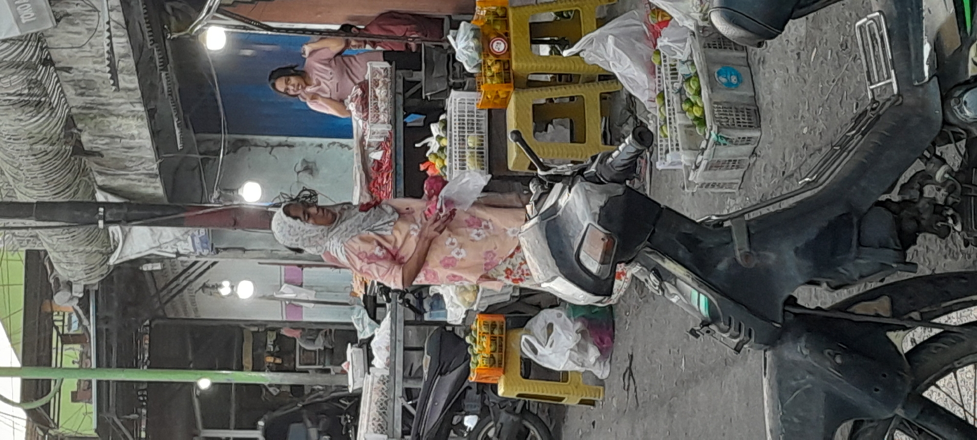 In Rogojampi, Indonesia, a street market scene unfolds with a focus on a woman in a floral-patterned dress and headscarf standing in the foreground. She holds a plastic bag in her right hand and appears to be interacting with a stall or examining produce. To her right, another woman in a pink shirt stands behind a market stall displaying what appears to be meat. The background reveals more market stalls, some with awnings, and a variety of goods, including fruits and vegetables in crates. Scooters and yellow plastic stools are visible around the market. The overall environment suggests a busy, open-air marketplace.